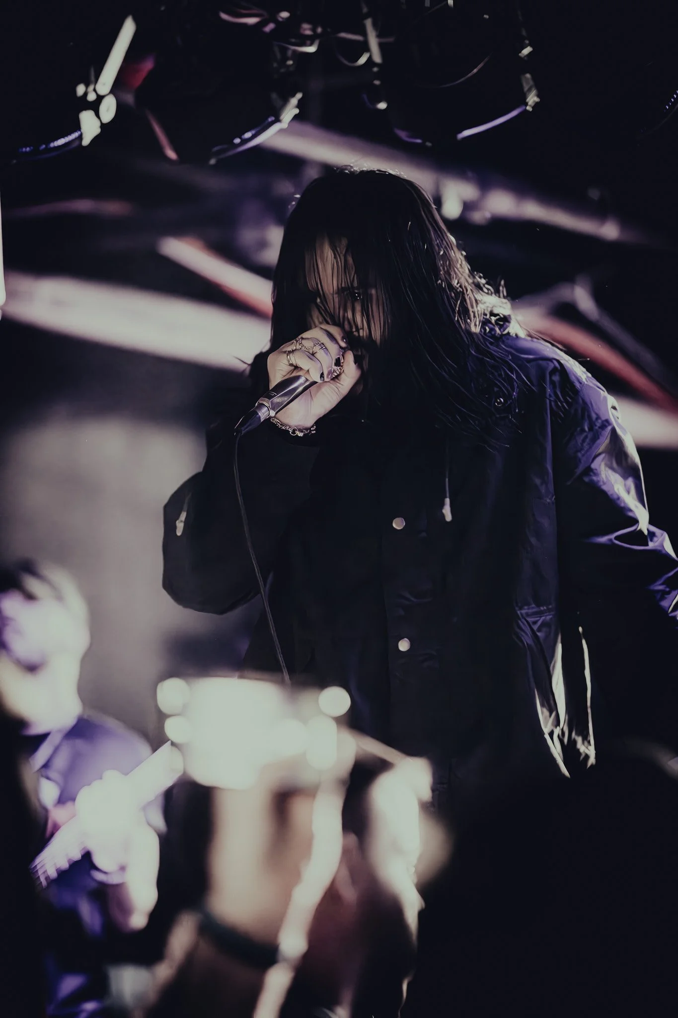 A singer with wet hair on stage, holding a microphone, with an audience member taking a photo in the foreground.