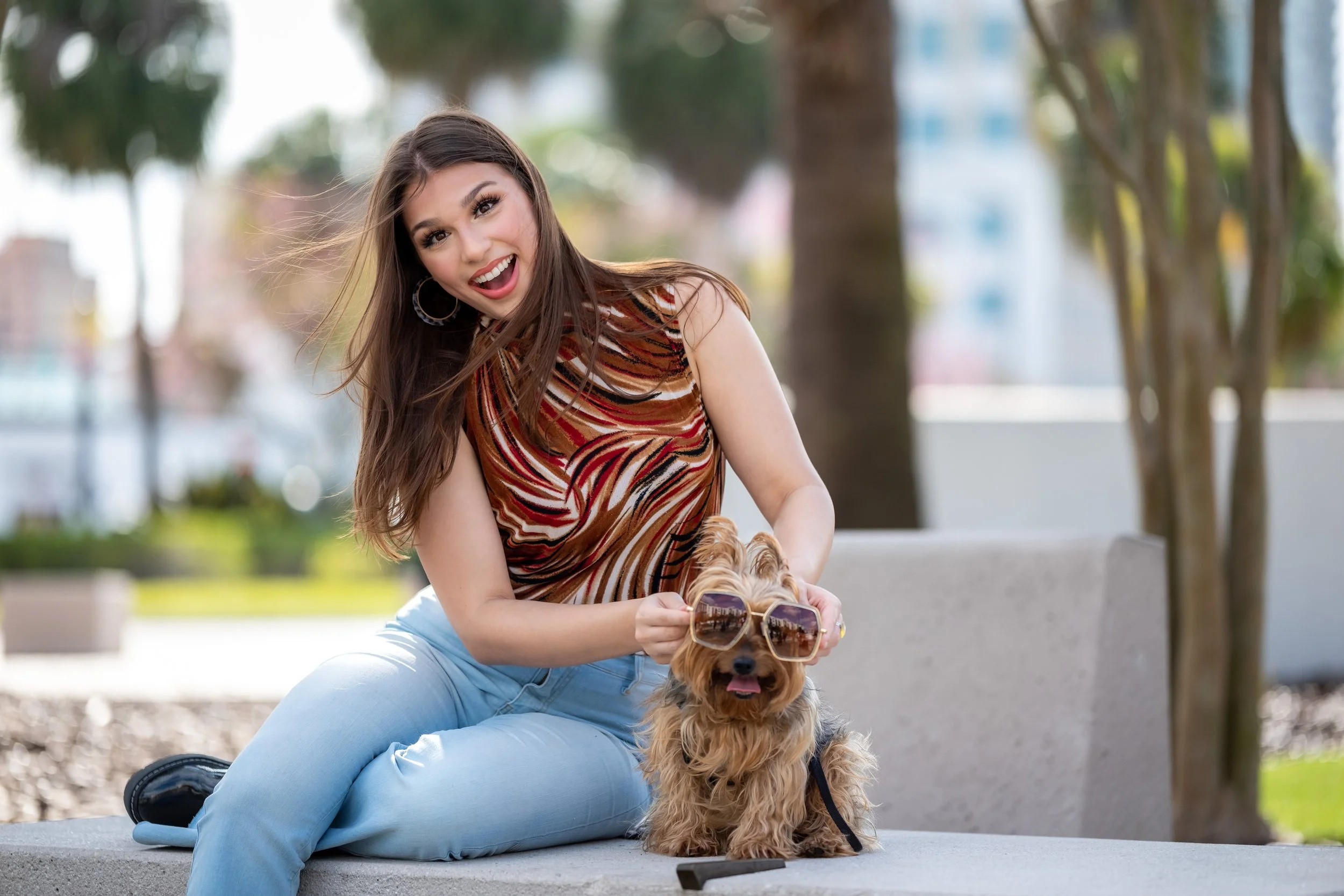 A young woman sitting on a bench outdoors wearing a colorful sleeveless top and light blue jeans, holding sunglasses over a small dog's eyes, with the dog sitting on the bench wearing sunglasses and looking happy.