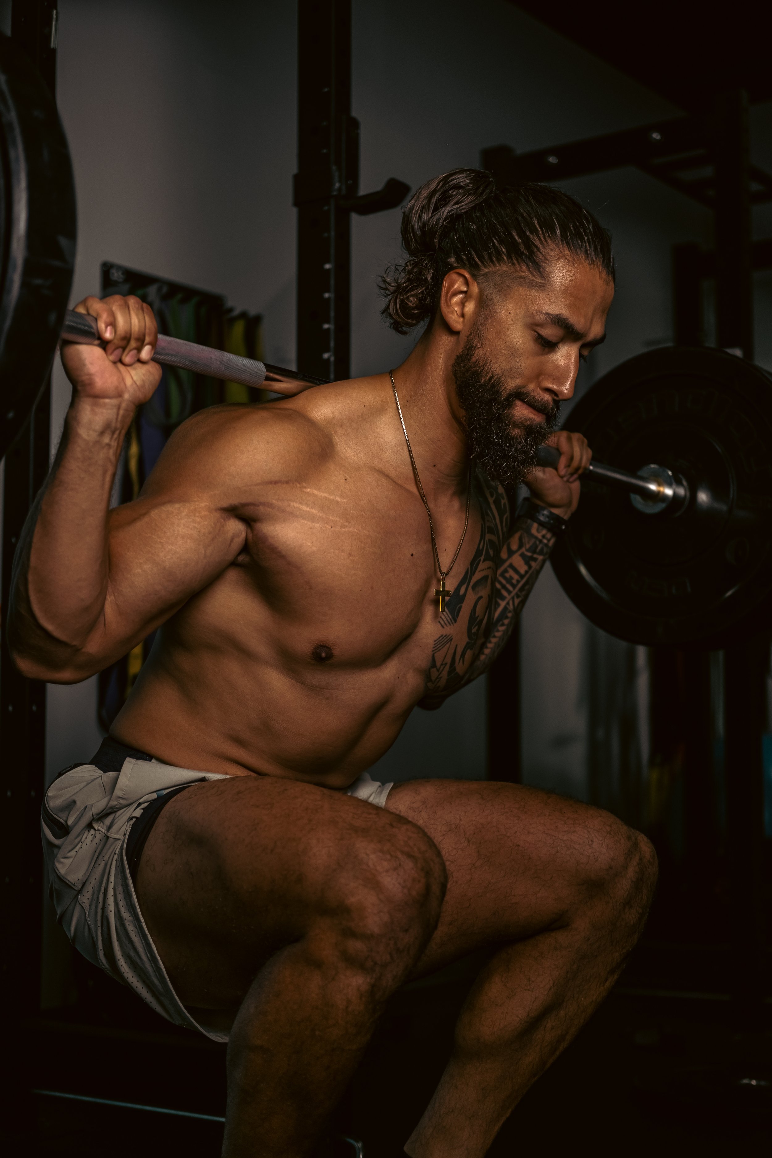 A shirtless man with a beard and tattoos doing squats with a barbell in a gym.