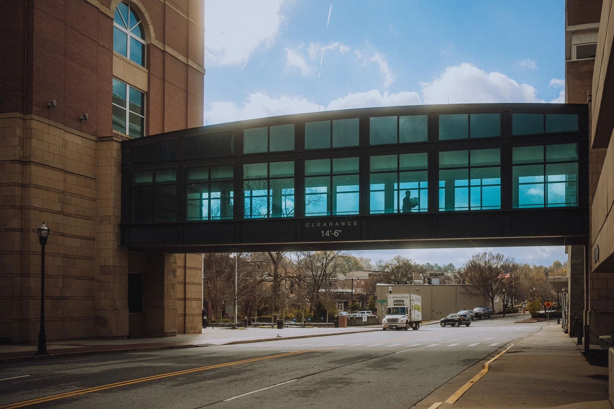 City street view featuring a skywalk bridge connecting two buildings, with a person visible inside taking a photo, daytime with partly cloudy sky, cars on the road, and leafless trees.