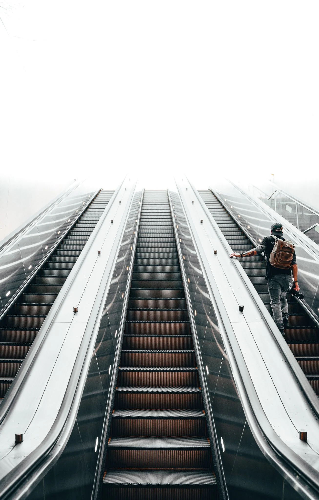 A person with a backpack riding an escalator in a modern, bright, and foggy indoor setting.
