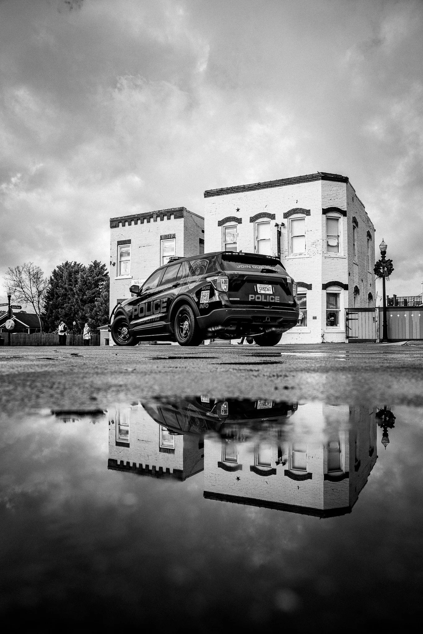 Black and white photo of a police SUV parked in front of a two-story brick building, with clouds in the sky and its reflection visible in a puddle on the ground.
