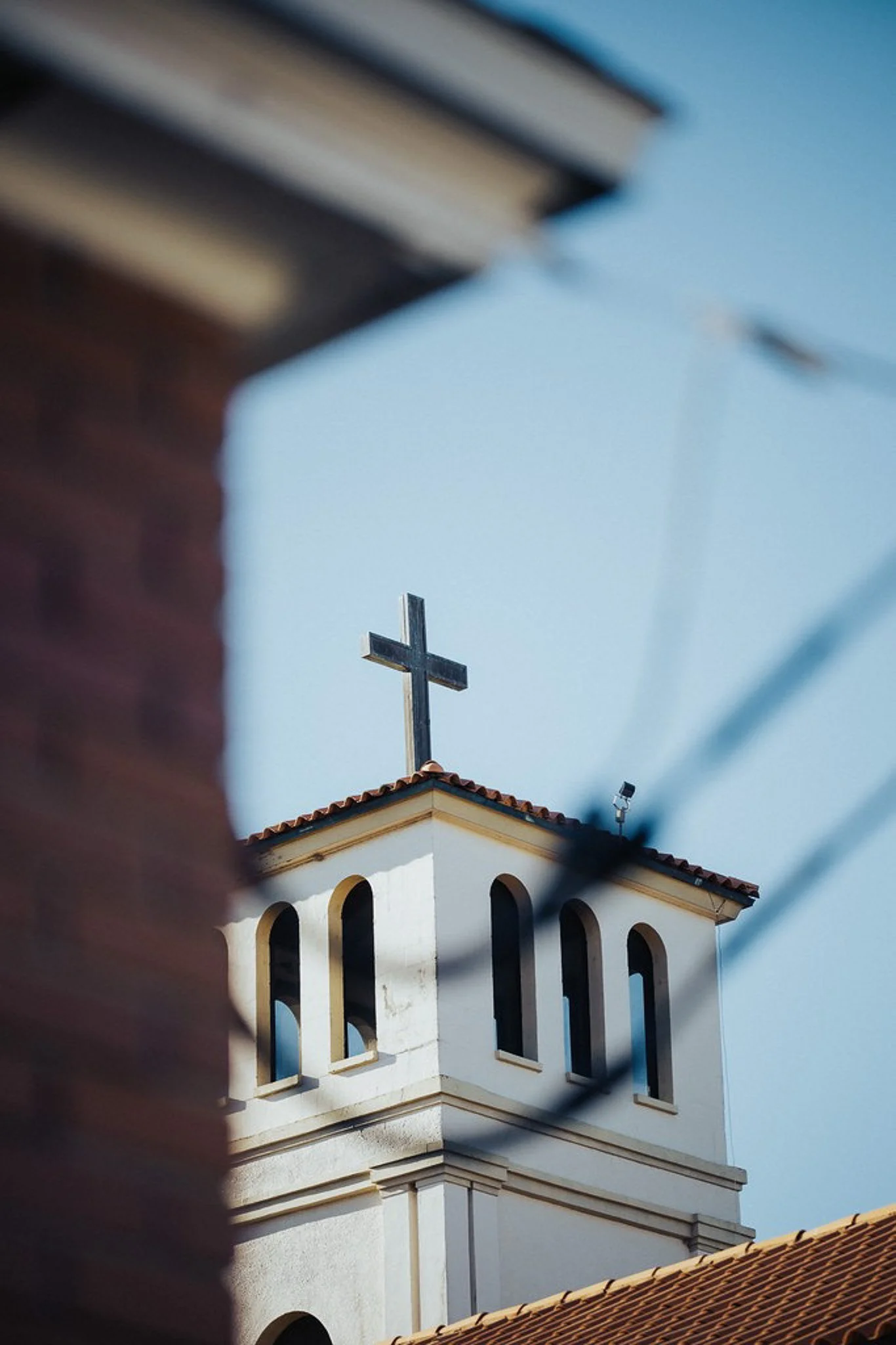 A church steeple with a cross on top, viewed through a blurred foreground of a brick wall and a black iron fence, under a clear blue sky.