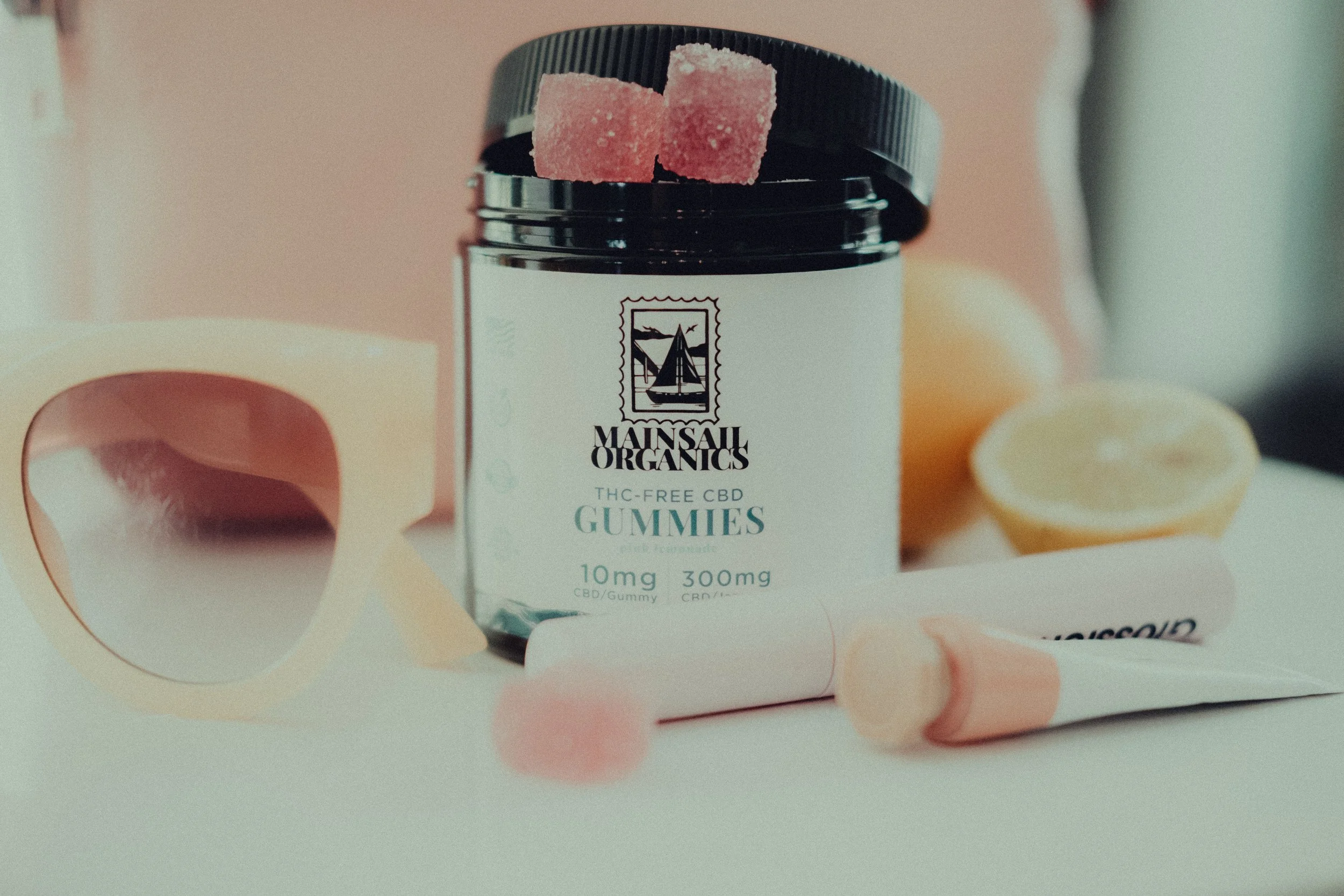 A jar of Mainsail Organics THC-free CBD gummies with gummies spilling out, surrounded by makeup-ware, a lemon, and a tube of lotion, on a white surface.
