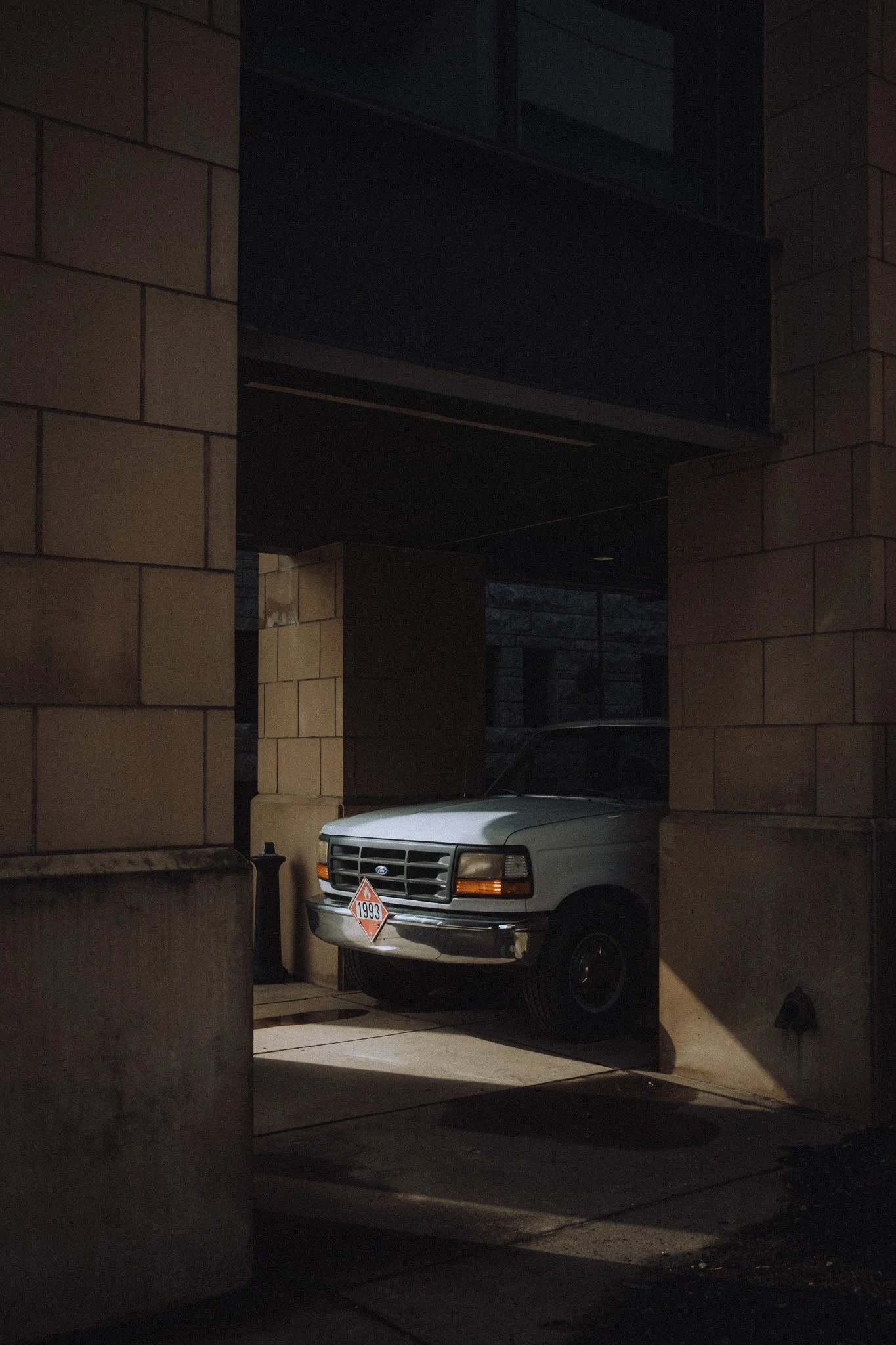 A white vintage car parked in a shadowy urban alleyway, with a 1993 hazardous material sign on the front.