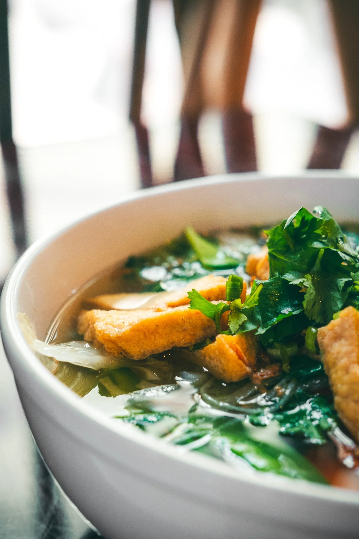 Close-up of a bowl of hot soup with vegetables, fried tofu, and fresh herbs, set on a table near wooden chairs.