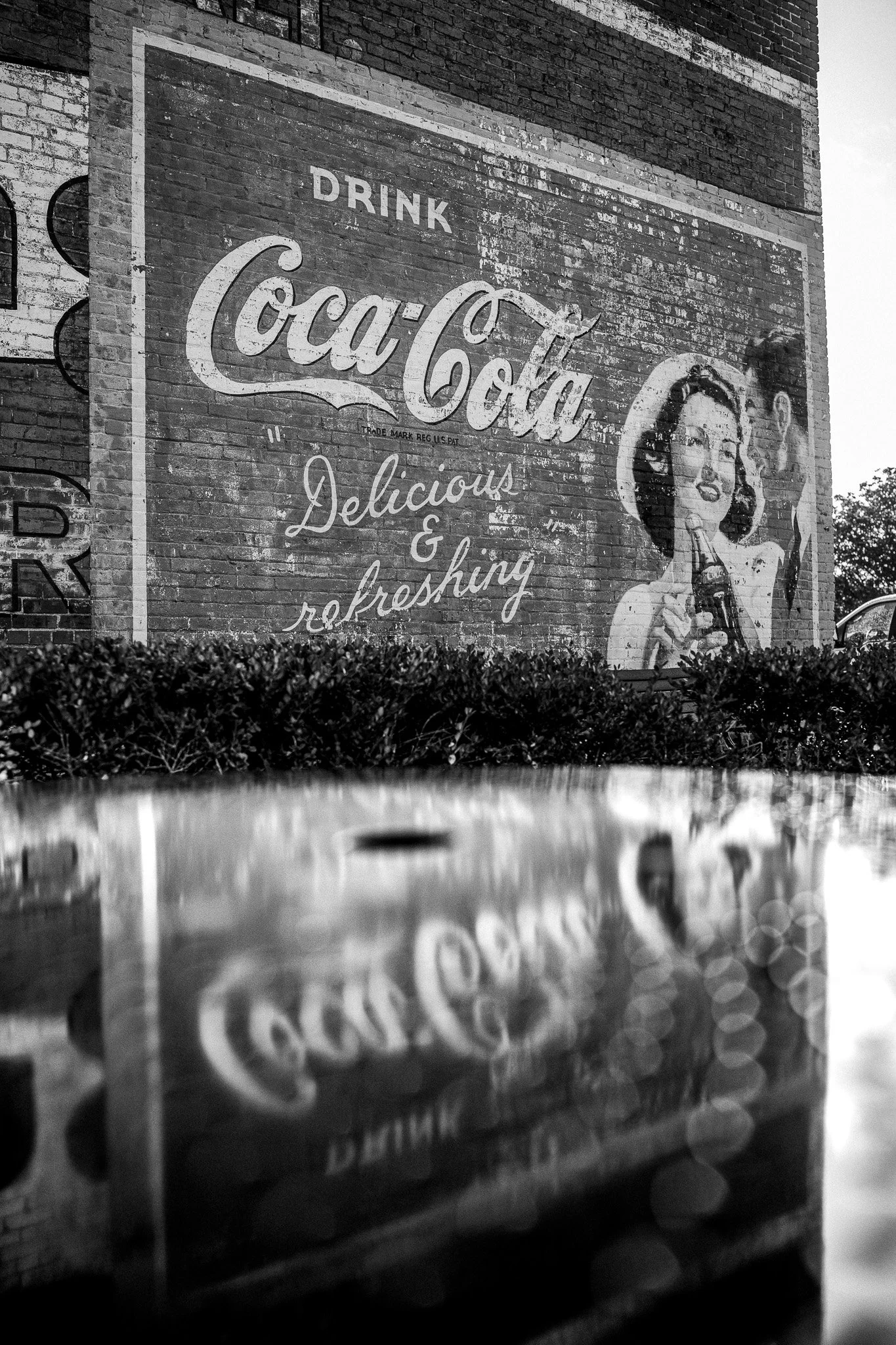 Black and white mural on brick wall advertising Coca-Cola, with a vintage style image of a woman holding a bottle and the words "Drink Coca-Cola, delicious & refreshing".