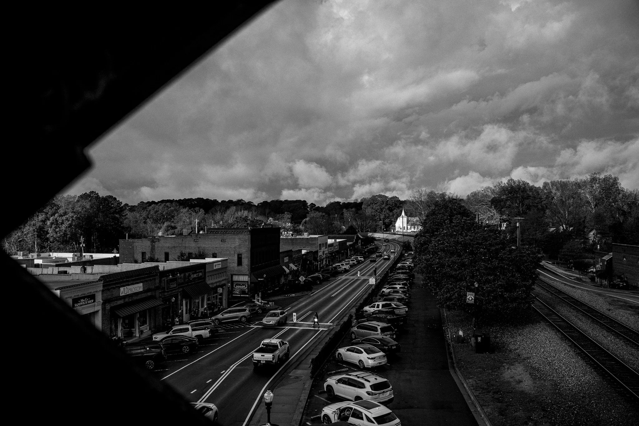 Black and white photo of a small town street with parked cars, a few people walking, businesses on the left, and train tracks on the right, under a cloudy sky.