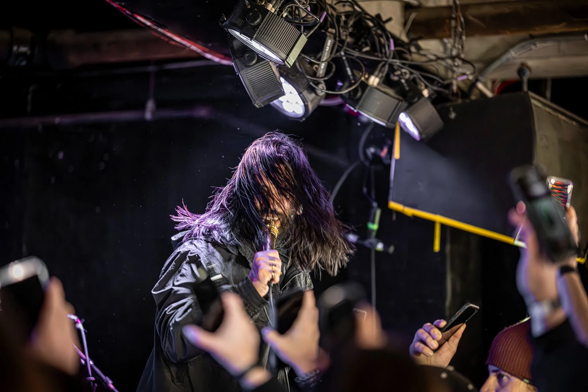 Female singer with long dark hair singing into a microphone on stage, surrounded by fans taking photos with their phones.