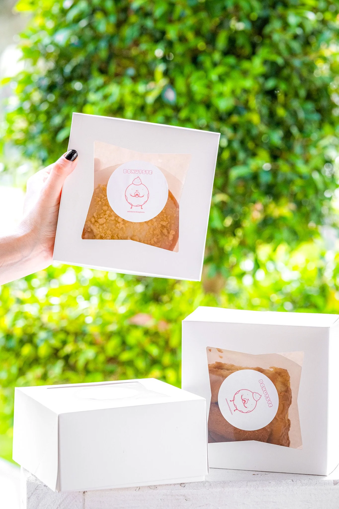 A person holding a box of donut treats in front of a lush green background, with two more donut boxes on a white surface.