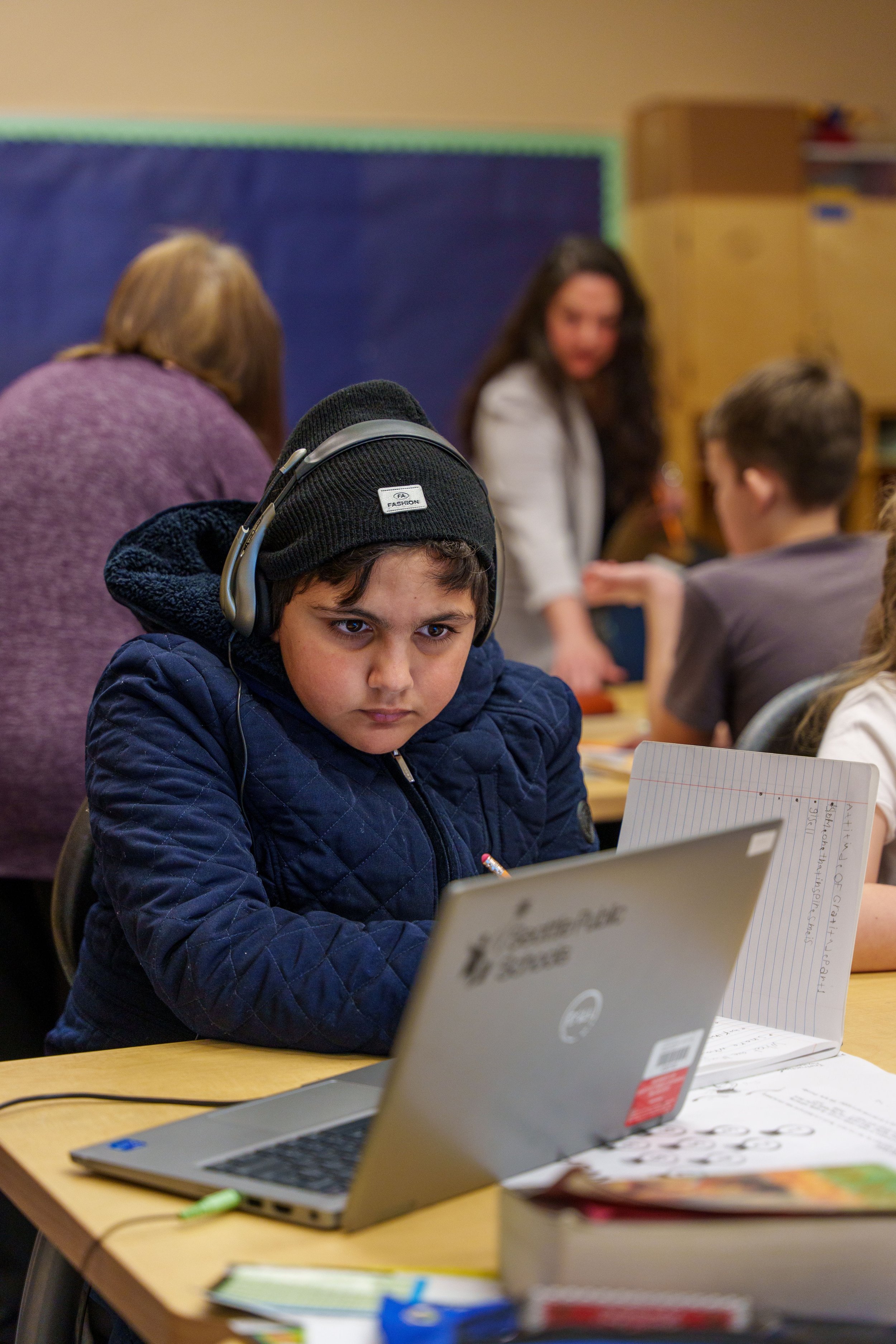 Boy with headphones peering at an open laptop. Sitting at a desk in a busy classroom.