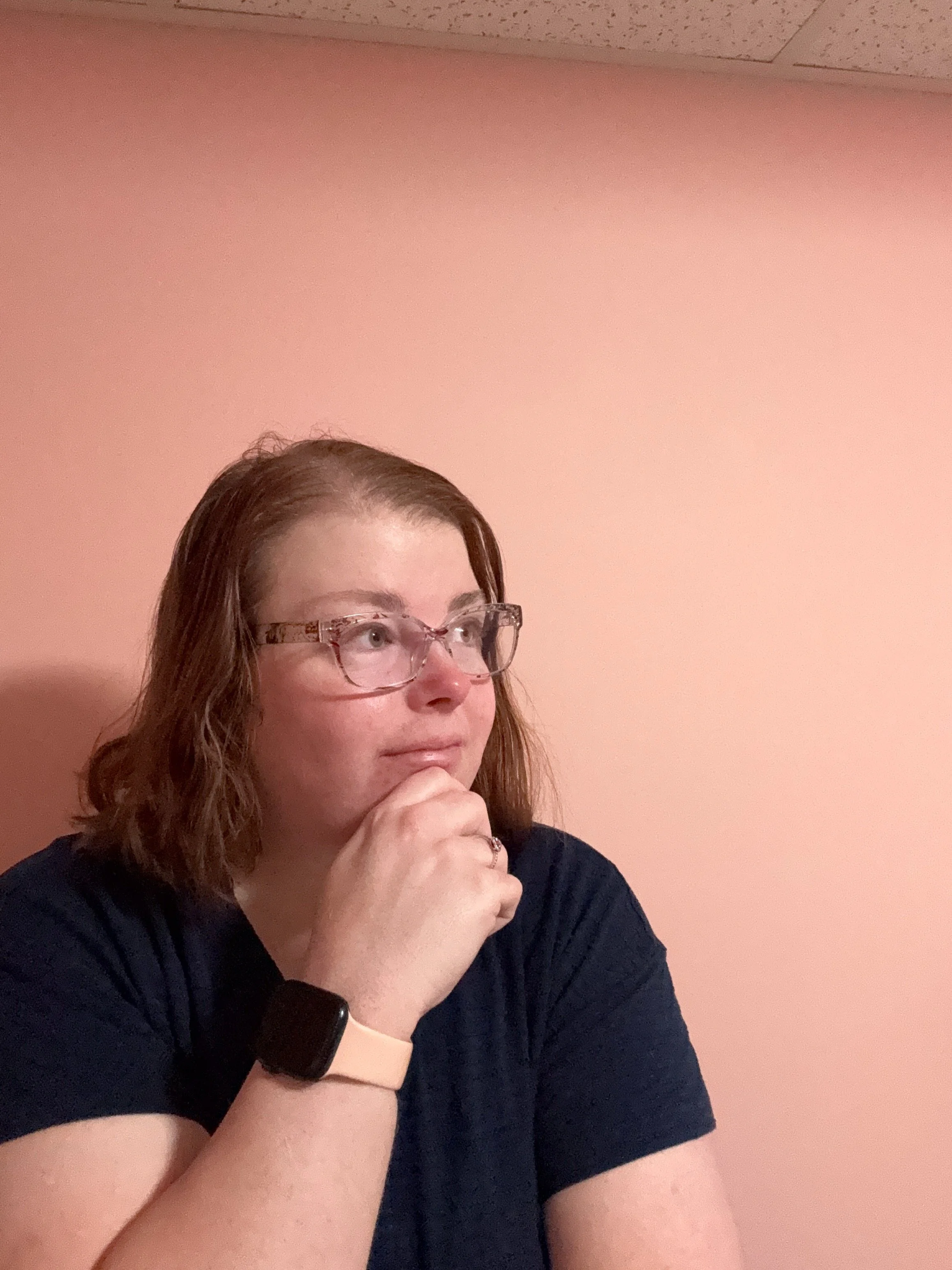 A woman with glasses and red hair, wearing a navy blue shirt, sitting against a pink wall, in a thoughtful pose with her chin resting on her hand.