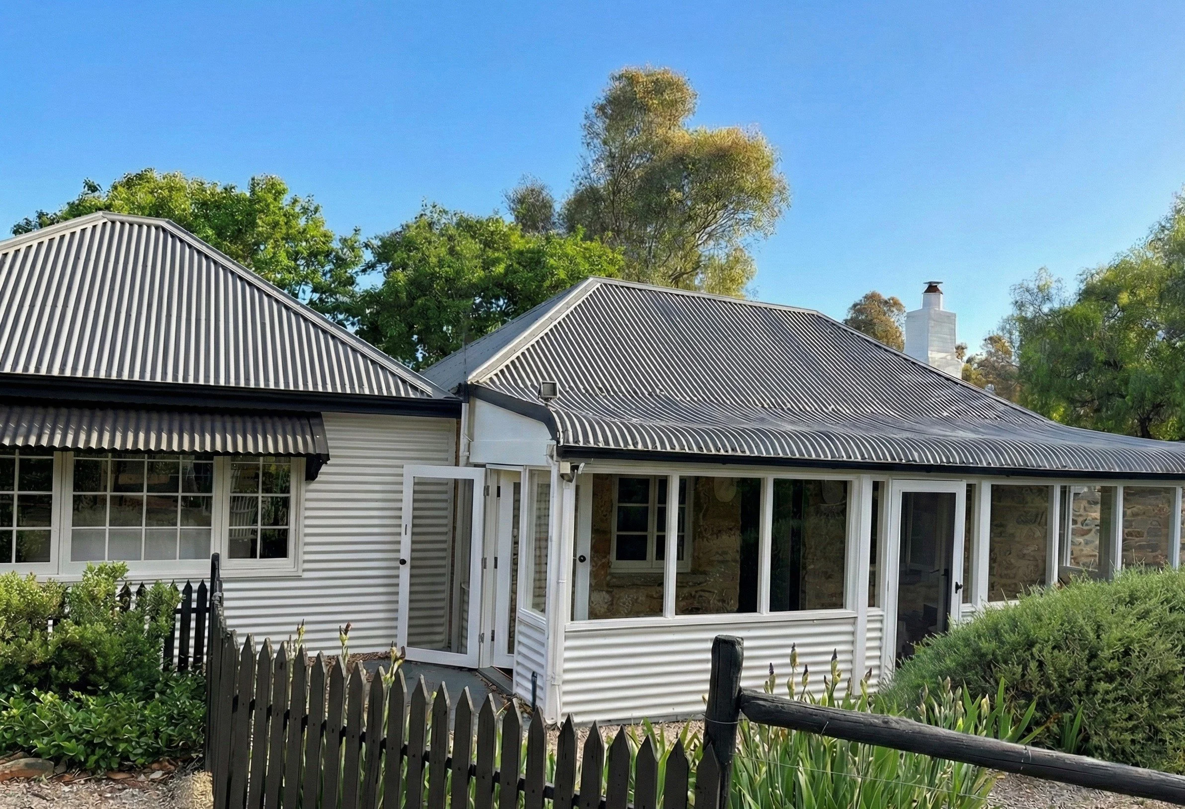 House with a corrugated metal roof, enclosed porch with glass windows, and a black picket fence, surrounded by green bushes and trees, under a clear blue sky.