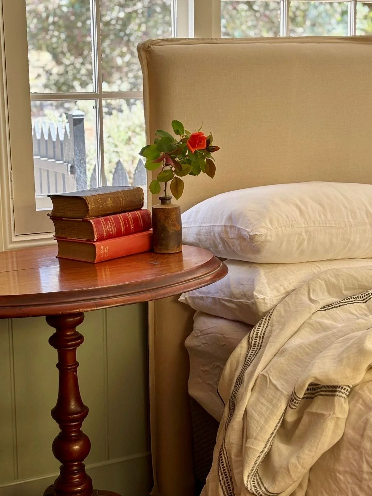 A bedside table with a small flower vase containing a red flower, three stacked books, a bed with white pillow and cream-colored bedding, and a window in the background showing outdoor foliage.