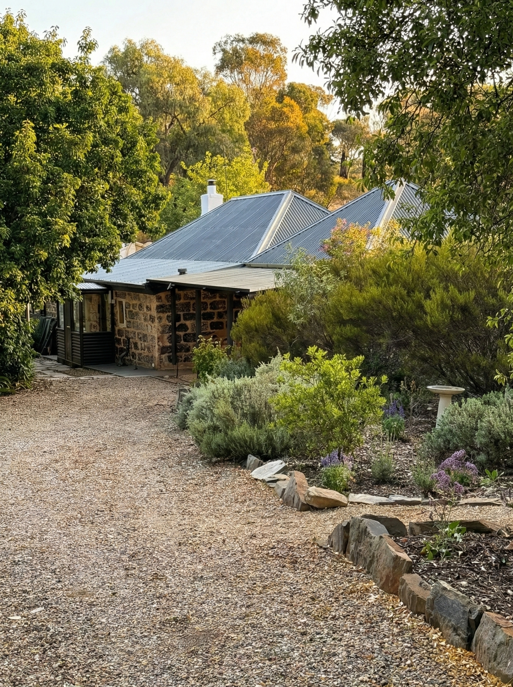 A rustic house with a stone exterior and corrugated metal roof, surrounded by lush greenery and a gravel pathway in a garden setting, with trees in the background.