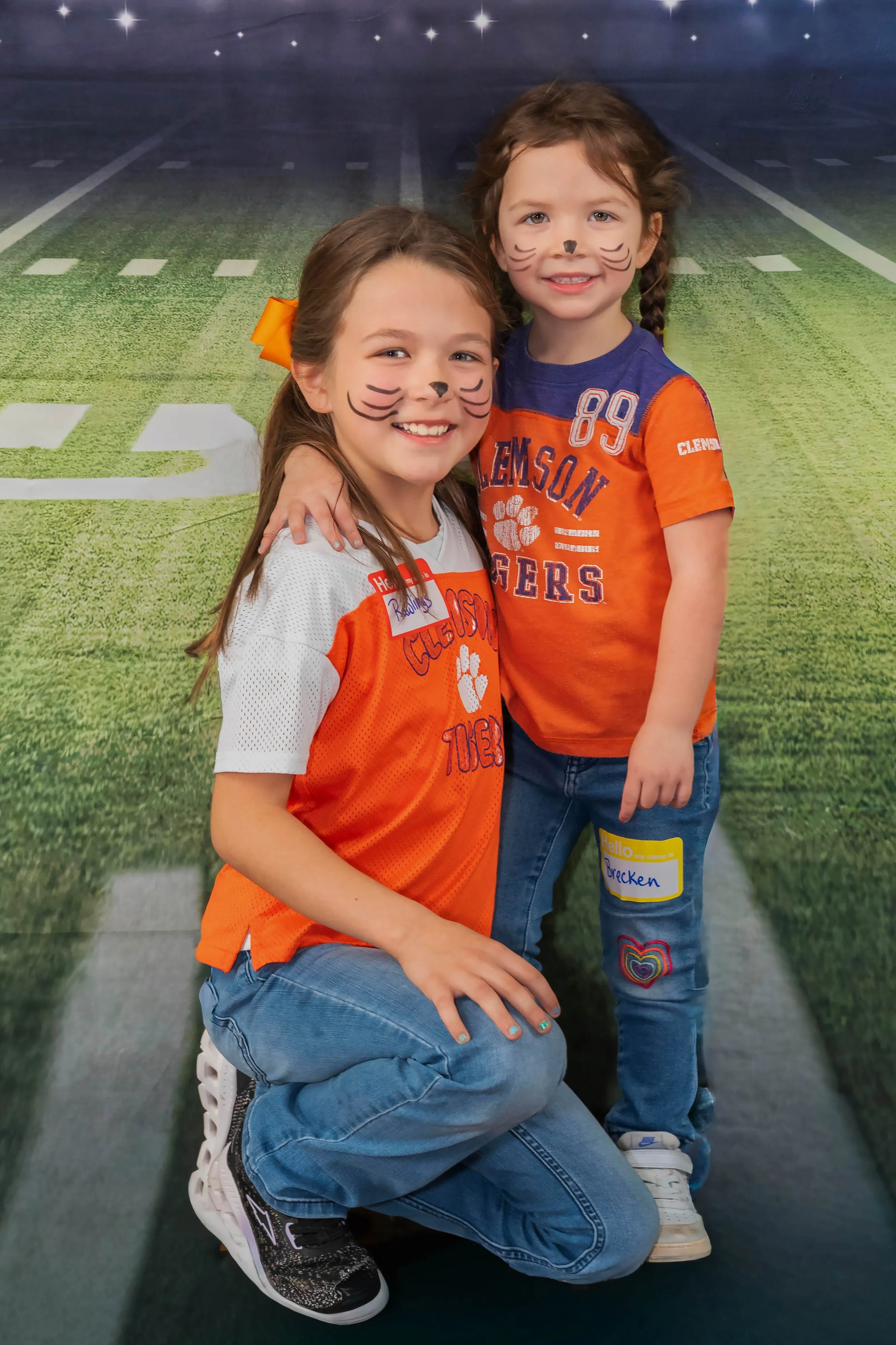 Two young girls with face paint resembling cats, wearing Clemson Tigers shirts, happily posing together on a football field background.