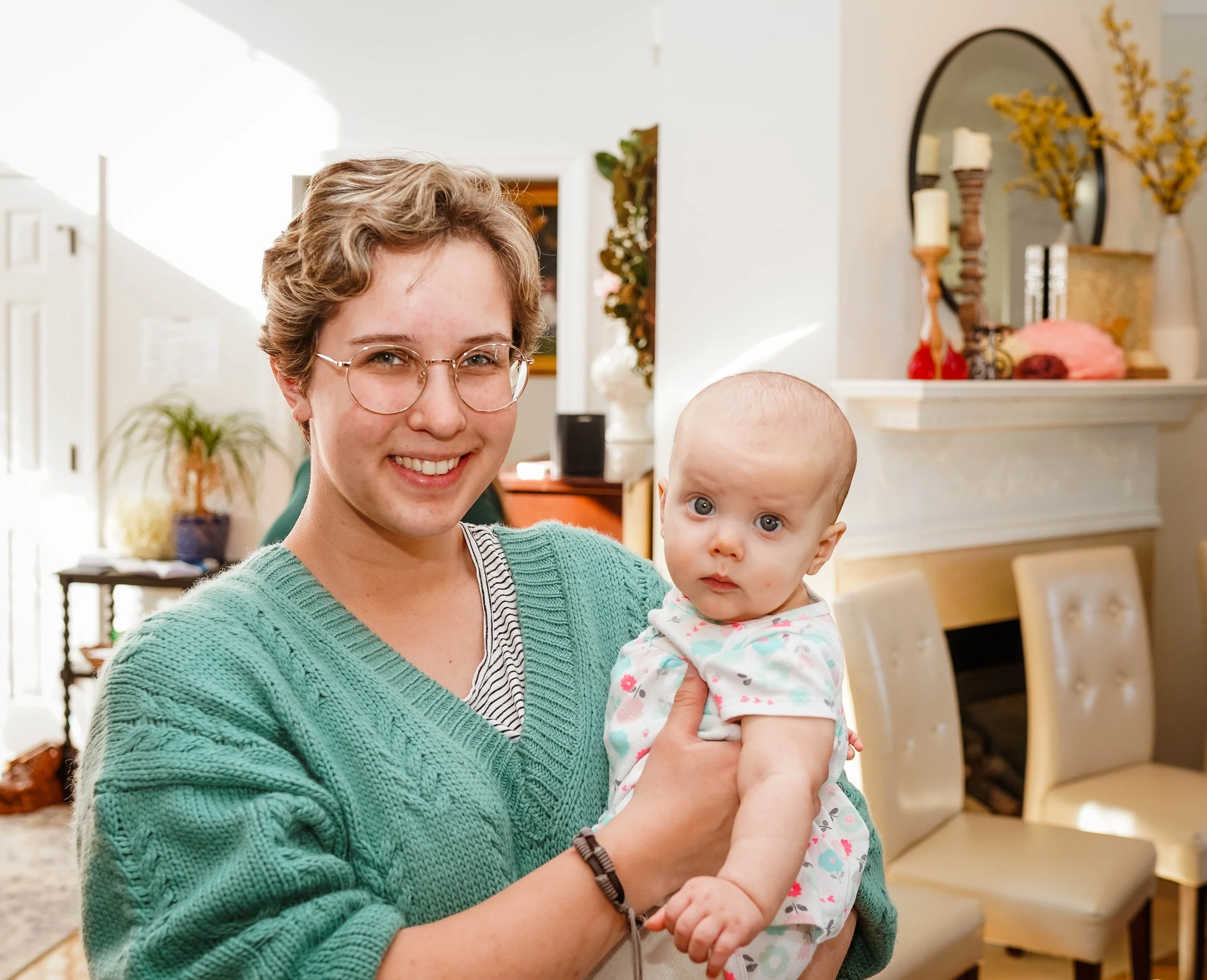 A young woman with short, curly hair, glasses, and a green sweater is smiling and holding a baby with a round face, big blue eyes, and wearing a white outfit with colorful polka dots inside a cozy home.