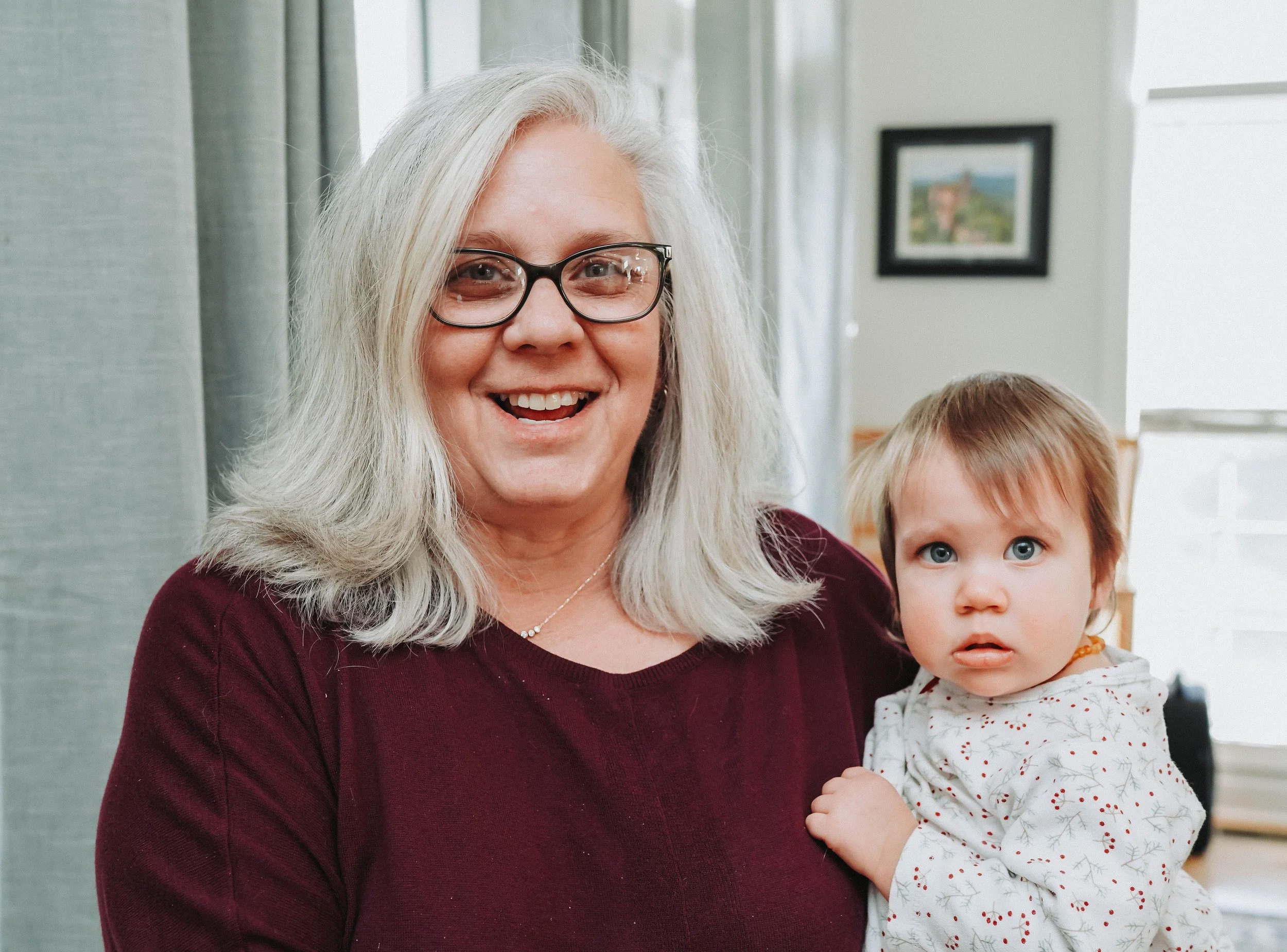 An elderly woman with gray hair, glasses, and a maroon top, smiling and holding a young girl with brown hair and blue eyes, wearing a holiday-patterned dress, inside a well-lit room with framed artwork on the wall.