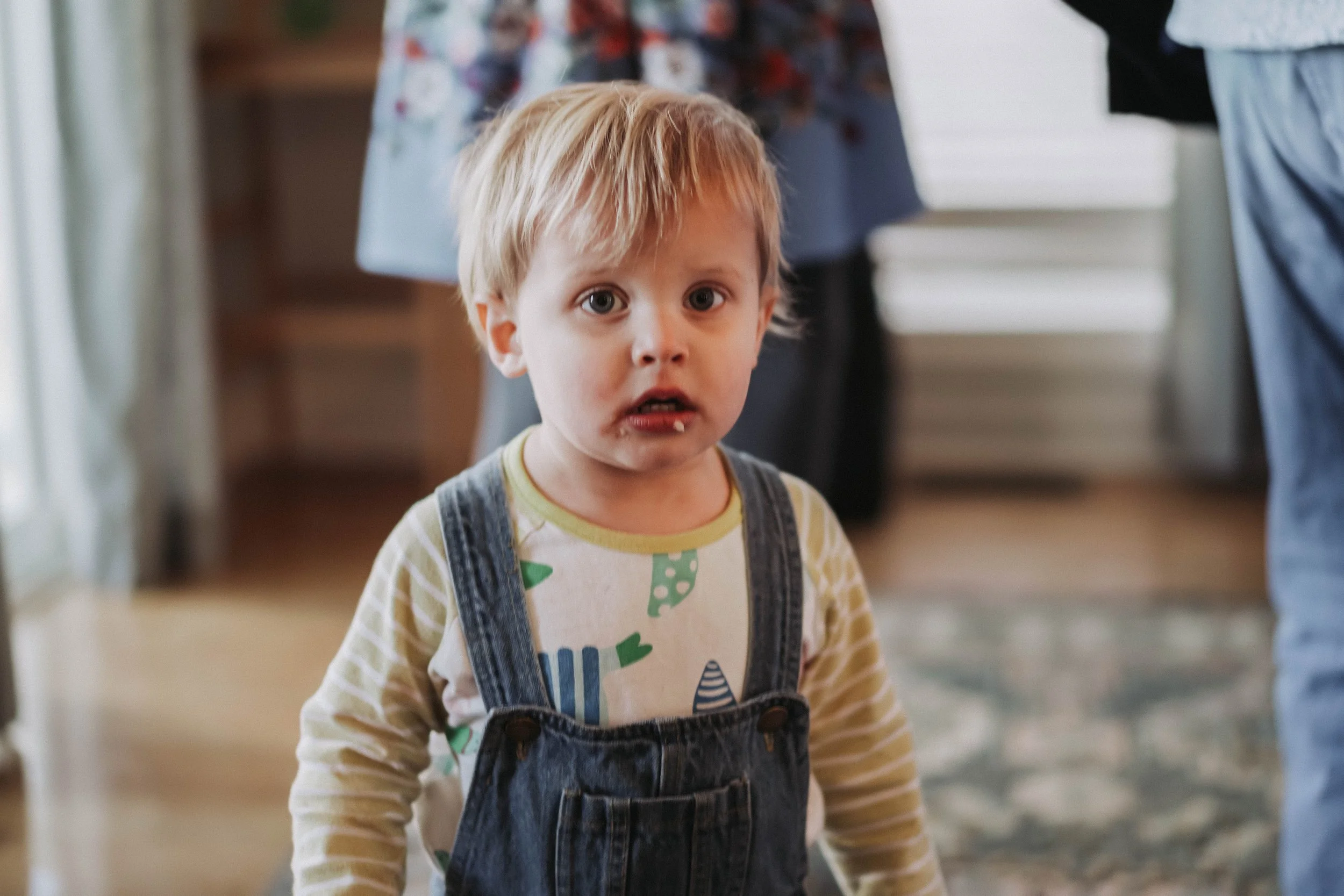A young boy with messy blonde hair and blue eyes looking directly at the camera, wearing a graphic long-sleeve shirt and denim overalls, standing indoors in front of a blurred background with people and furniture.
