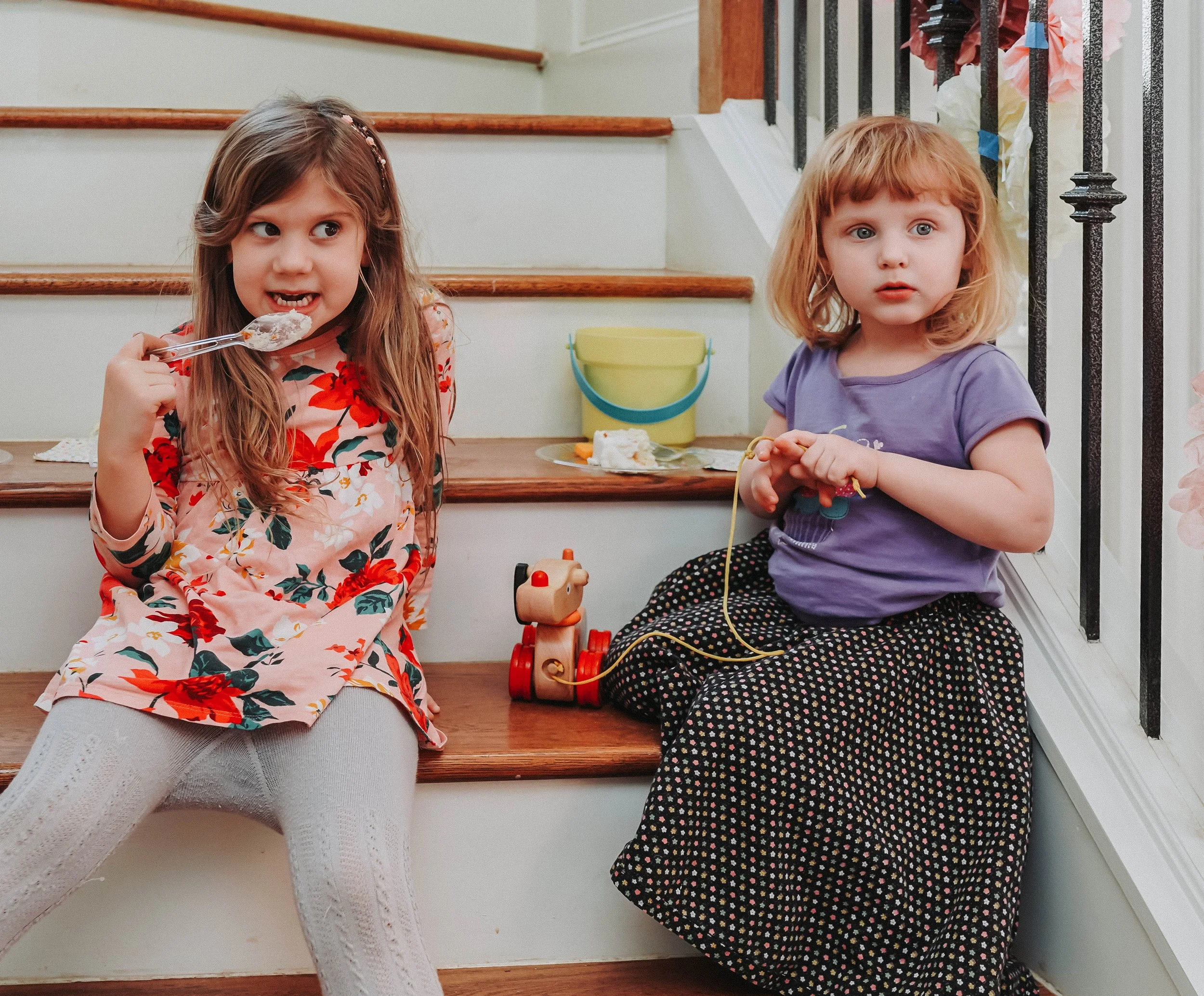 Two young girls sitting on wooden stairs indoors, one with long brown hair wearing a floral dress and white tights eating with a spoon, and the other with red hair wearing a purple shirt and patterned skirt holding a toy, with a toy dog nearby and a yellow bucket on the stairs.