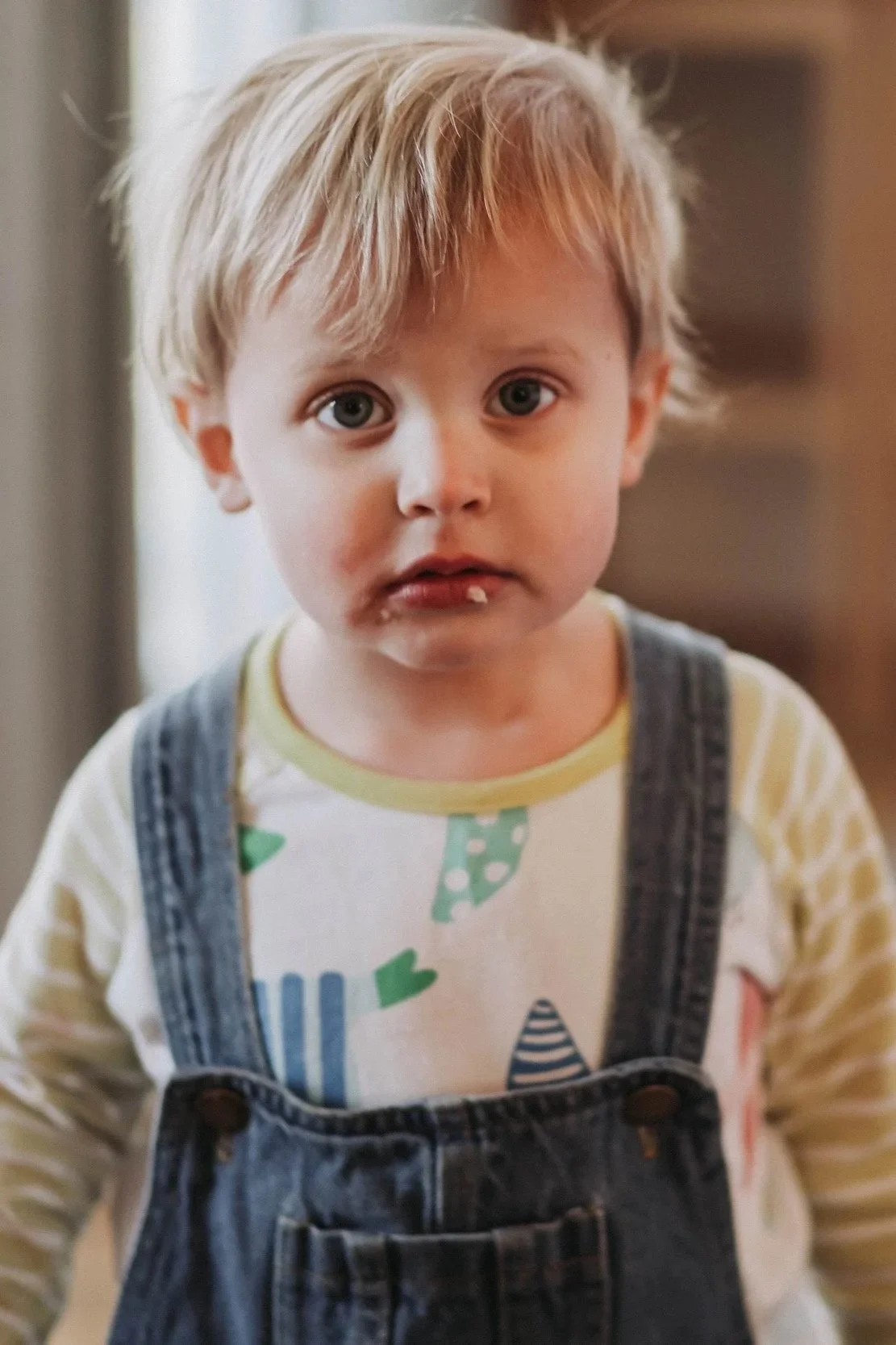 A young boy with blond hair and blue eyes looking directly at the camera, wearing a white shirt with colorful patterns and denim overalls, with food around his mouth.
