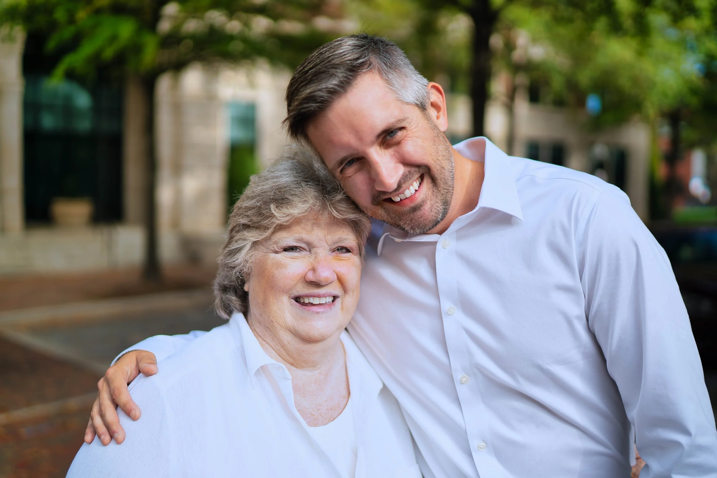 A middle-aged woman and a young man smiling and hugging outdoors, with trees and a building in the background.