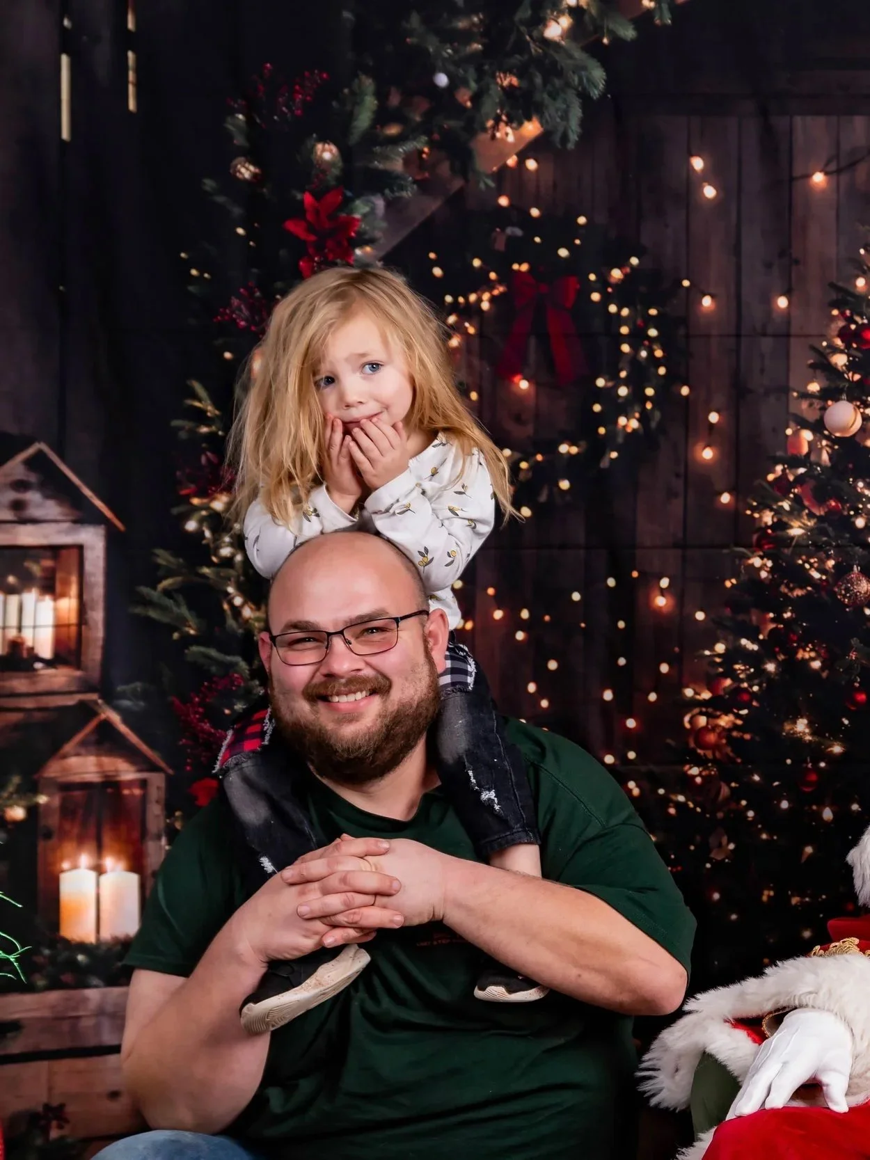A man with glasses and a beard smiling, with a young girl with red hair and a white shirt sitting on his shoulders, in front of Christmas decorations including a Christmas tree with ornaments and lights, and wreaths with red ribbons.