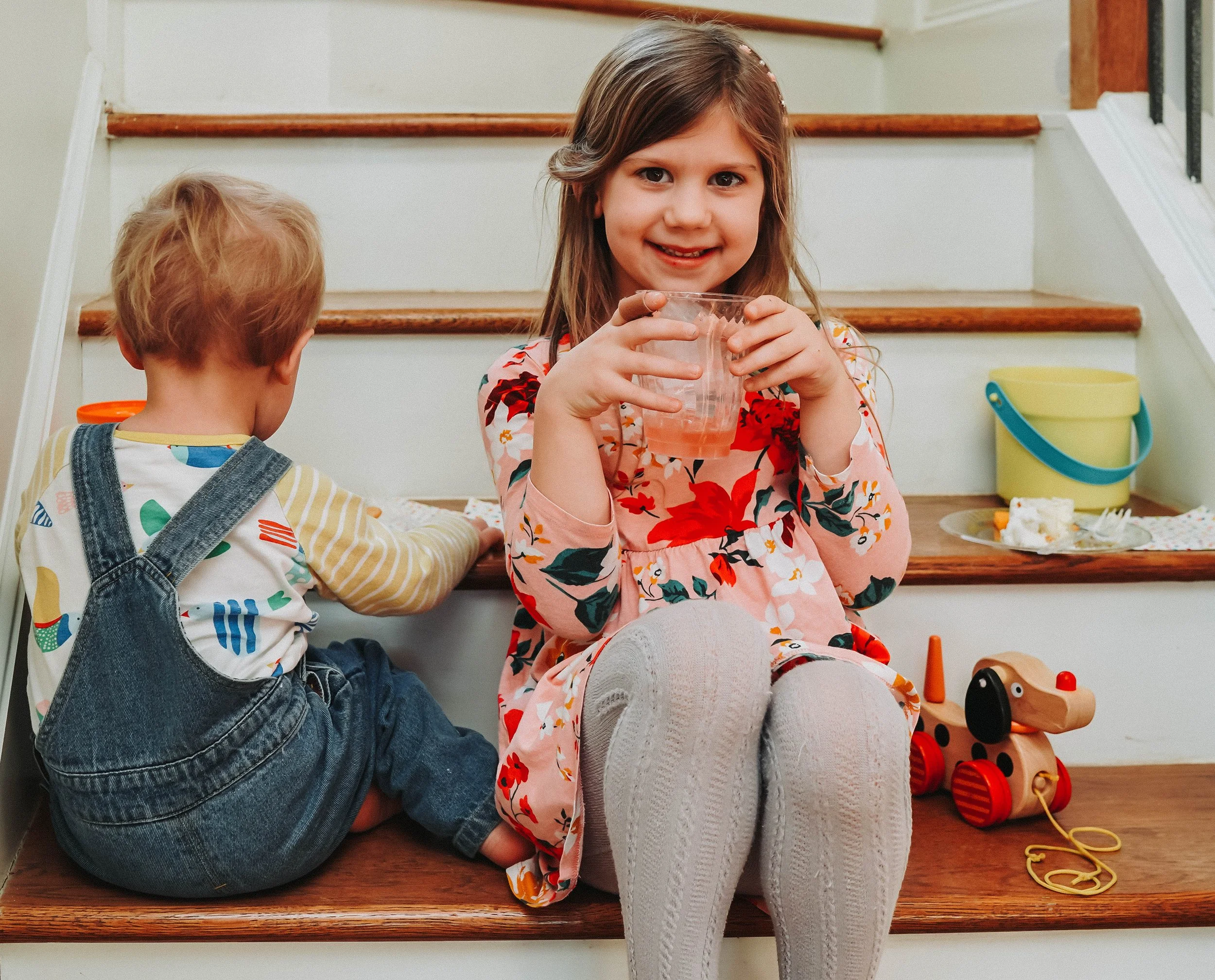 A young girl with long brown hair sitting on staircase, smiling and holding a glass of drink, with a young boy with blond hair sitting beside her, playing on the stairs with toys and food in the background.