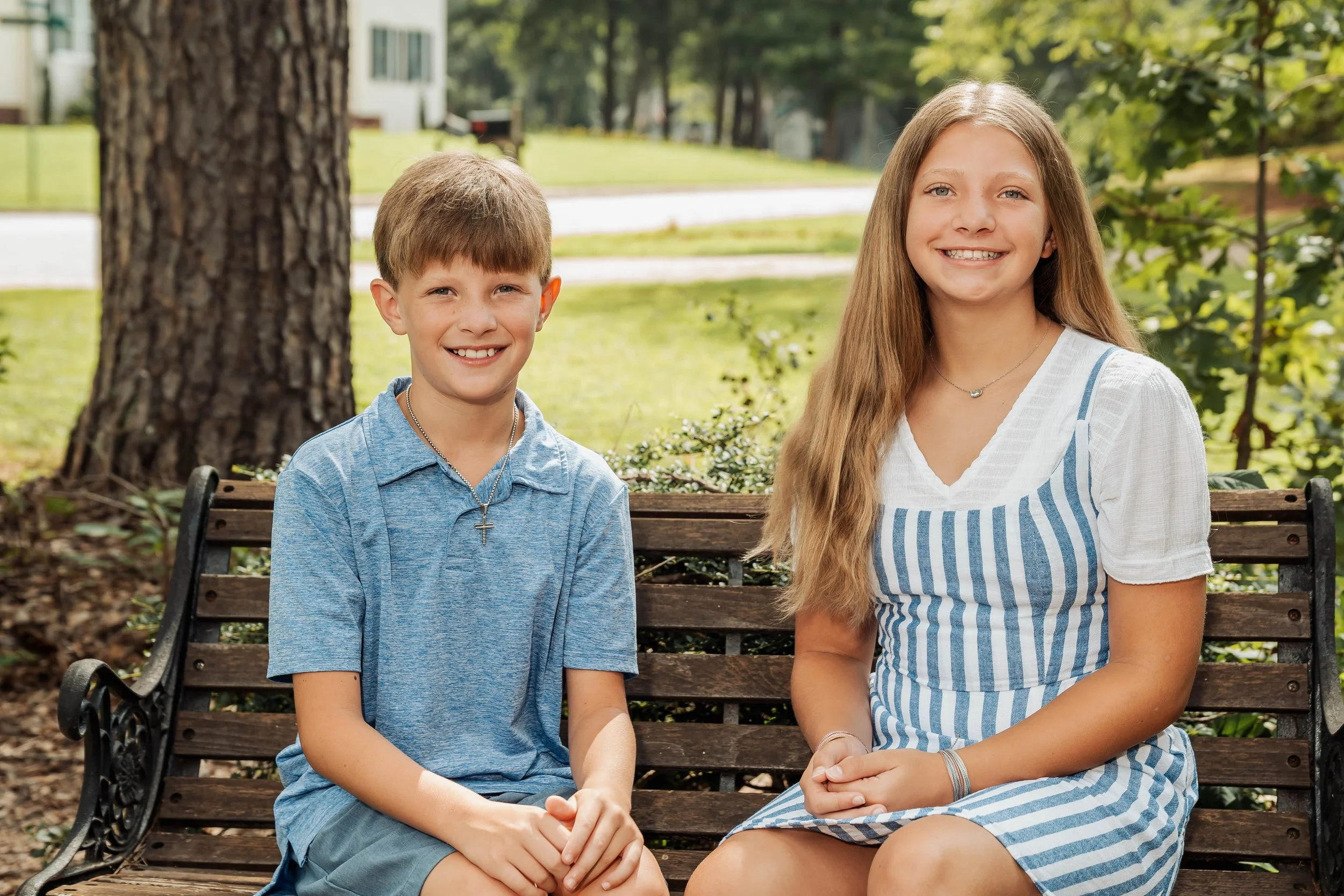 A young boy and girl sitting on a wooden park bench outdoors, smiling at the camera, with trees and greenery in the background.