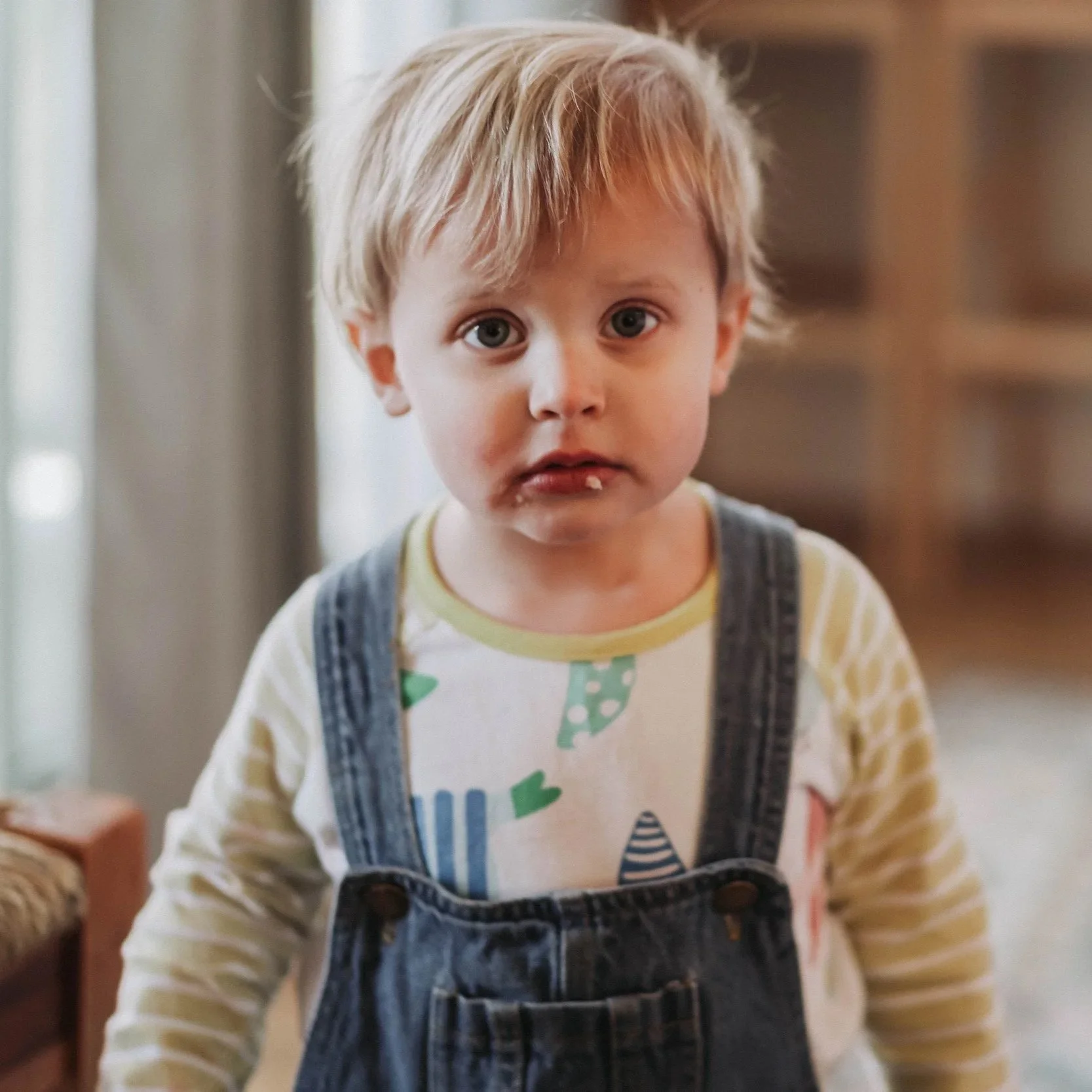 A young boy with blonde hair and blue eyes standing indoors, wearing a white shirt with a colorful dinosaur pattern and denim overalls.