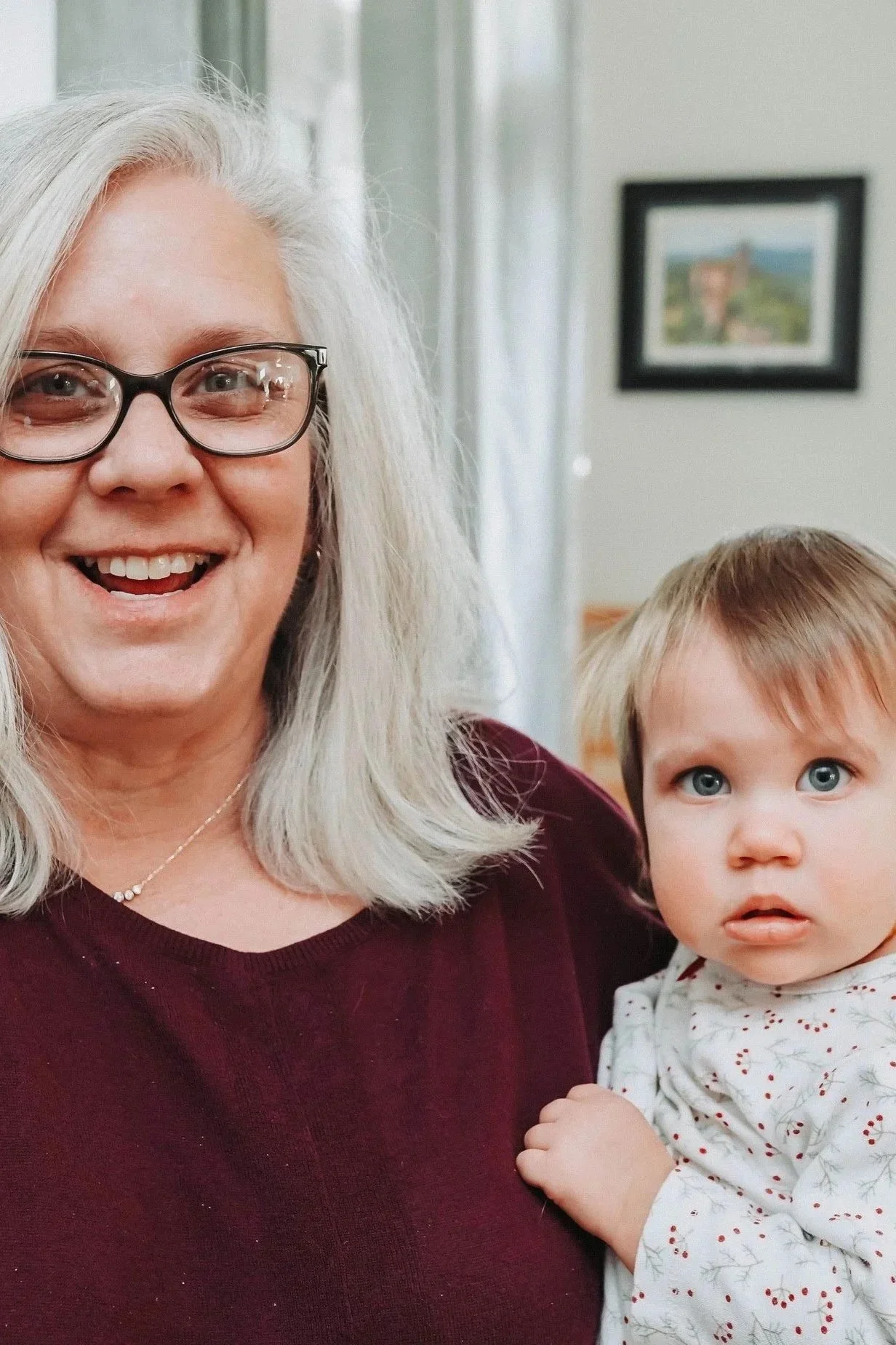 A woman with gray hair and glasses smiling at the camera, holding a young child with blue eyes and light brown hair, in a room with a framed picture on the wall in the background.