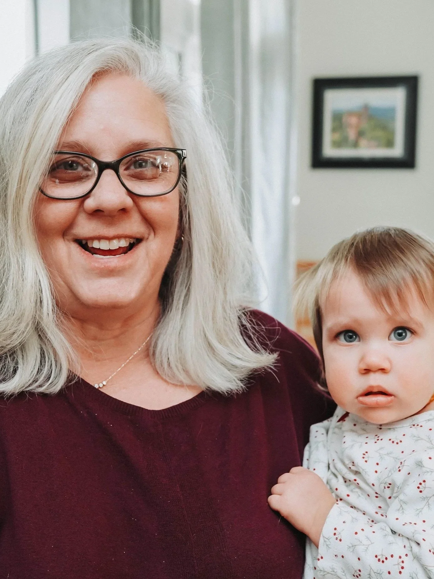 A smiling older woman with gray hair and glasses holding a young girl with blue eyes and light brown hair inside a home.