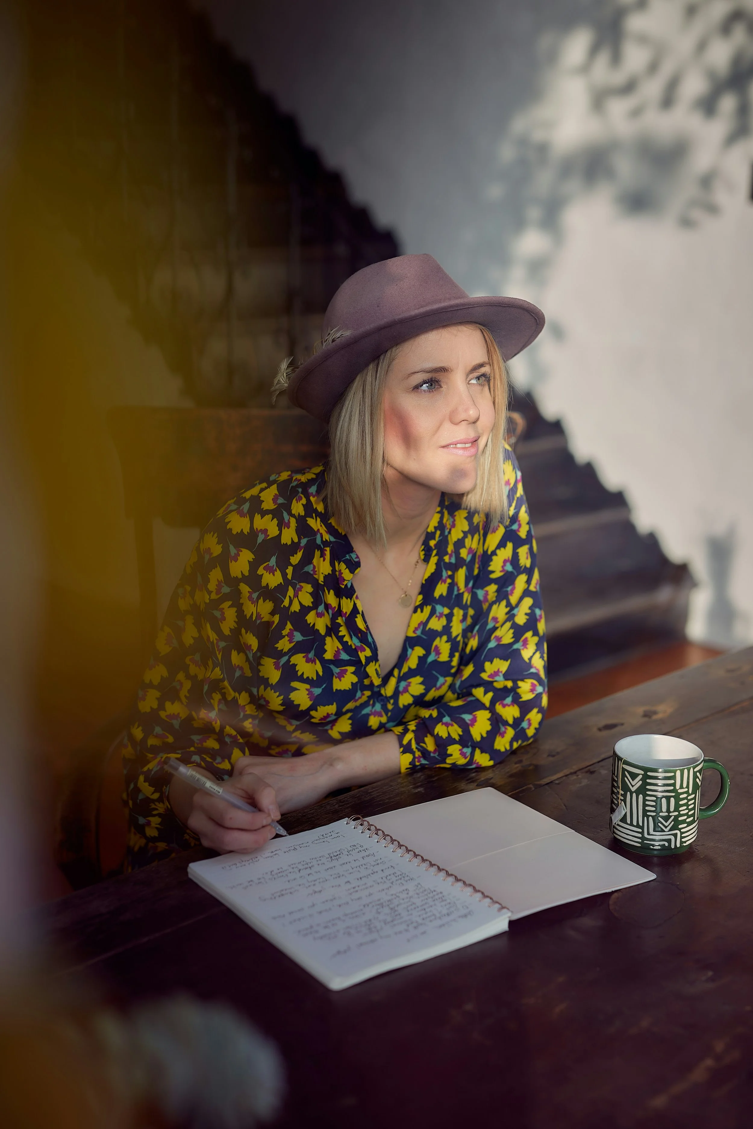 A woman in a colorful floral shirt and purple hat sits at a wooden table with a notebook and a green mug, looking thoughtfully to the side, with a blurred background of stairs and outdoor scenery.