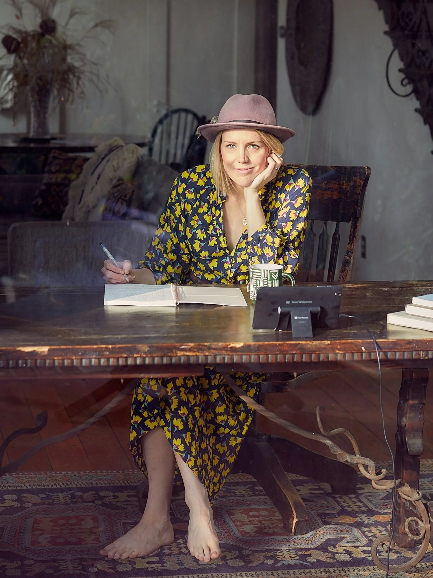 A woman sitting at a wooden table, viewed through a window, with notes, a pen, a mug, and a digital device in front of her. She is wearing a patterned dress and a wide-brimmed hat.
