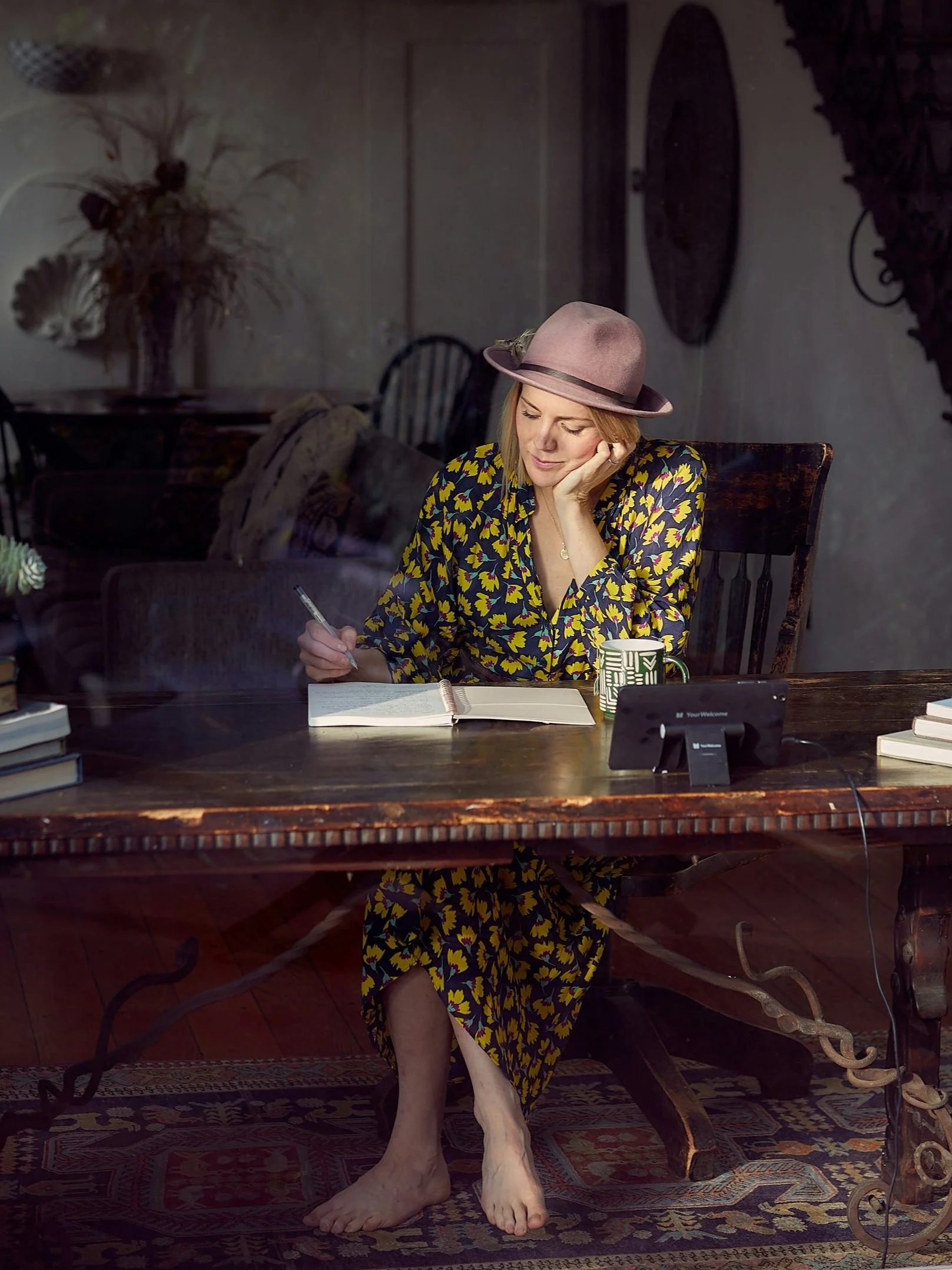 A woman in a colorful floral dress and a light pink hat, sitting at a wooden table, looking down and writing in a notebook, with a coffee mug, a tablet, and books nearby, inside a cozy room.
