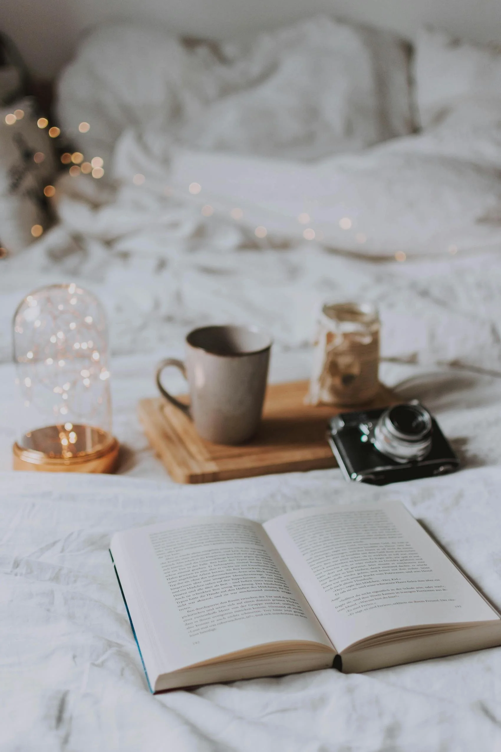 Open book on white bed with coffee mug, camera, jar, and string lights.