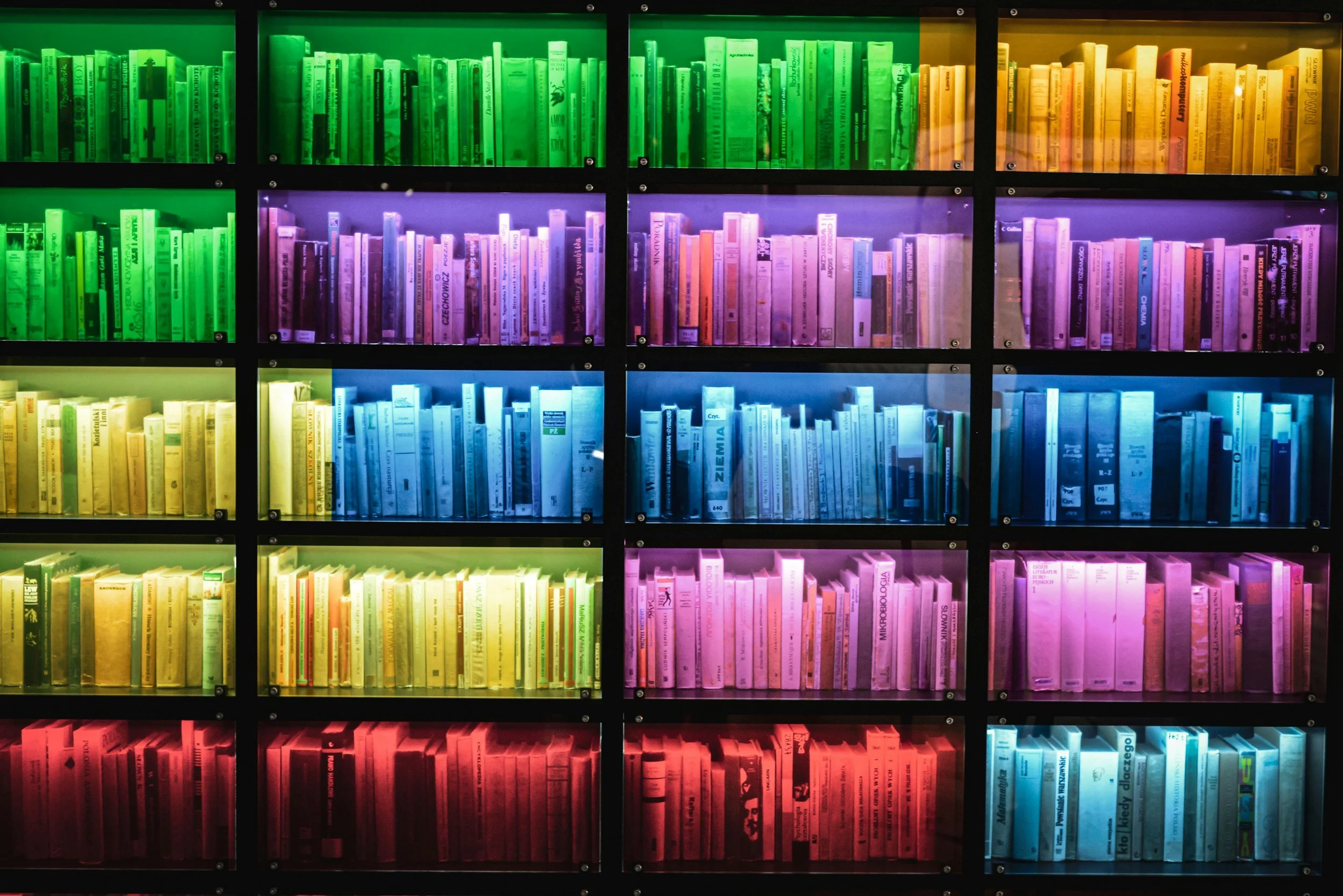 Colorful bookshelf with books illuminated by different colored lights, arranged in horizontal rows of red, yellow, green, blue, purple, and pink.