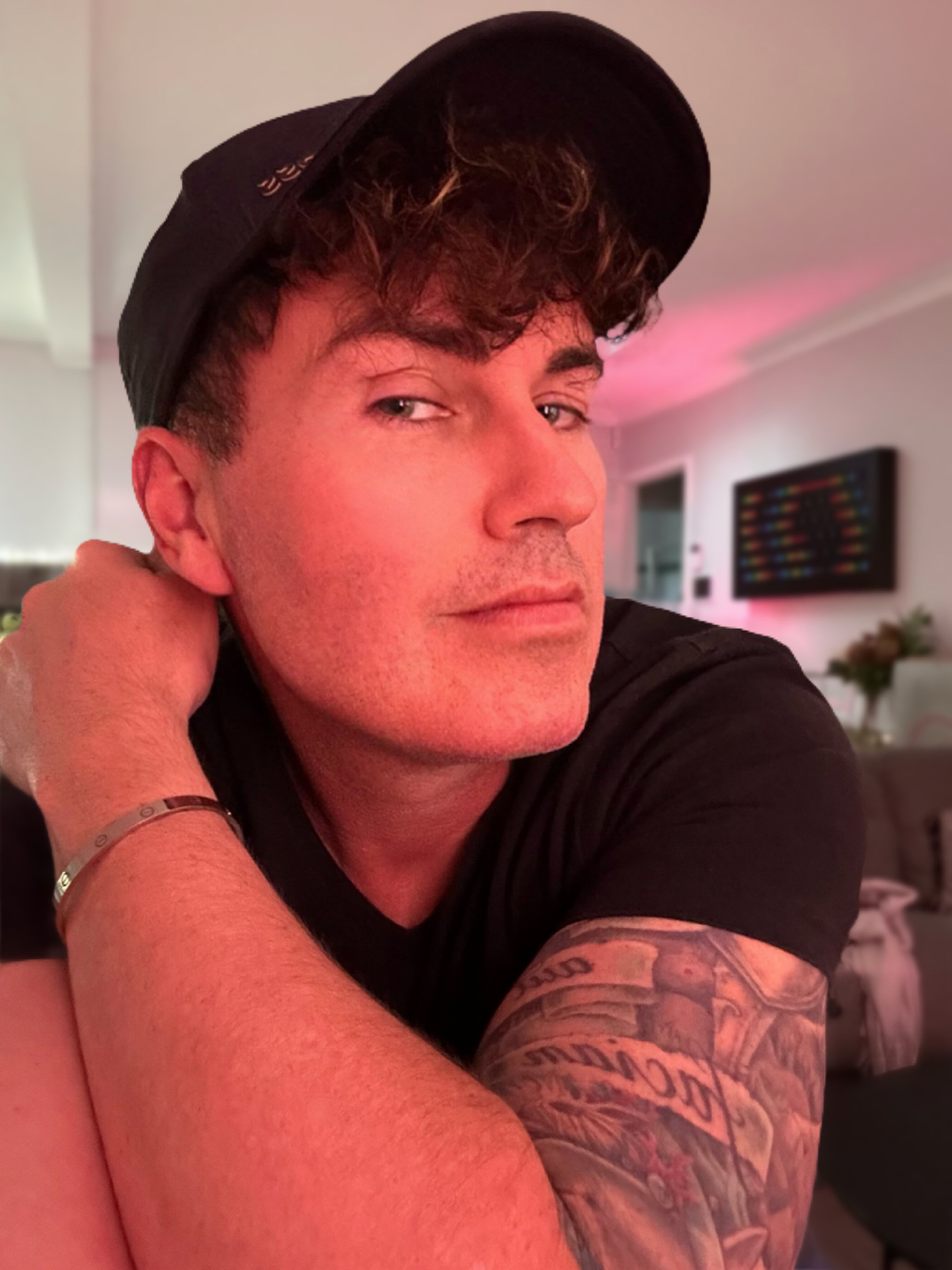 A young man with curly hair wearing a black cap and black shirt, resting his chin on his arm, in a cozy indoor setting with pink and purple lighting, a TV screen, and a sofa in the background.