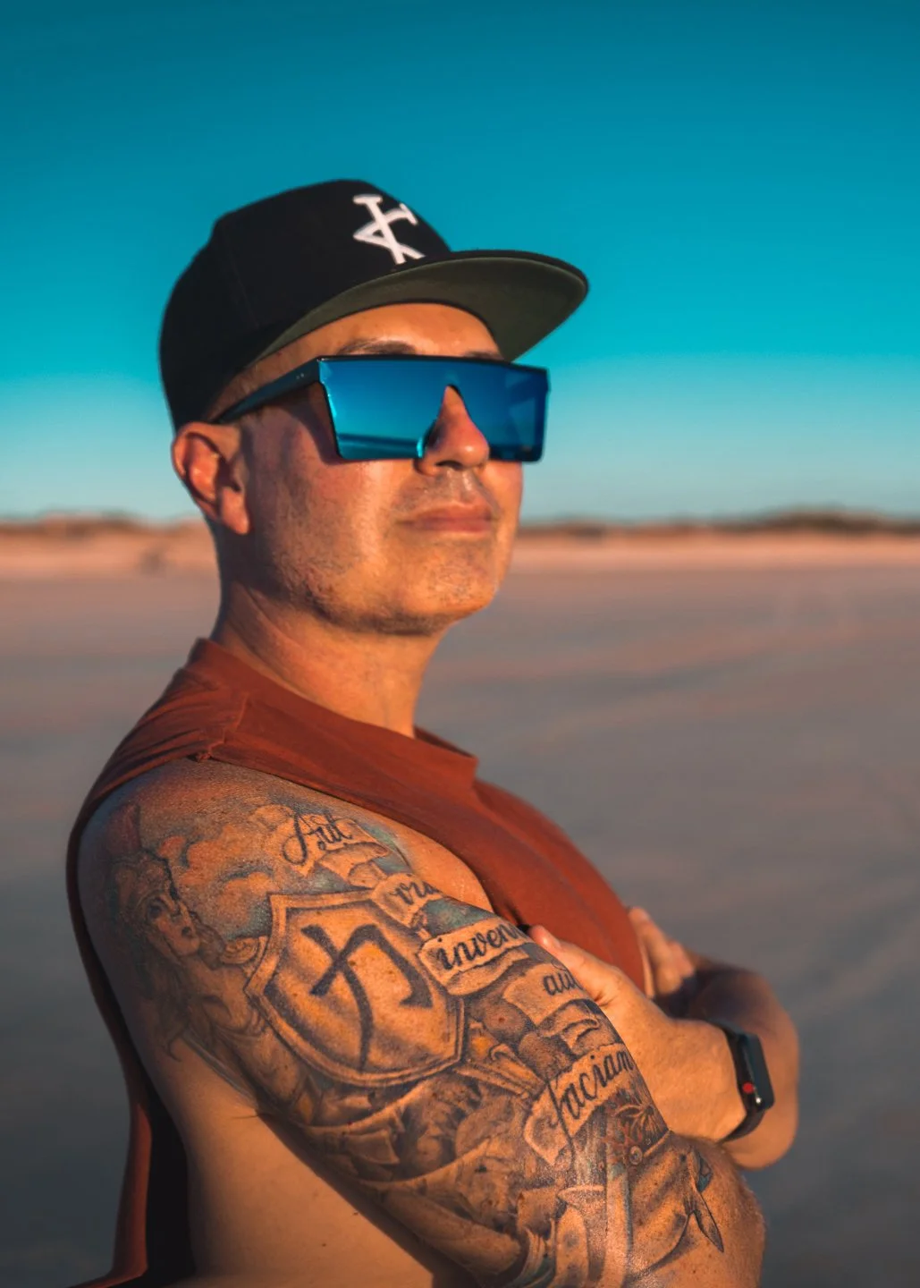 Man with tattoos on arm, wearing large blue sunglasses, a black baseball cap with white logo, and a sleeveless rust-colored shirt, standing on a sunny beach with arms crossed.