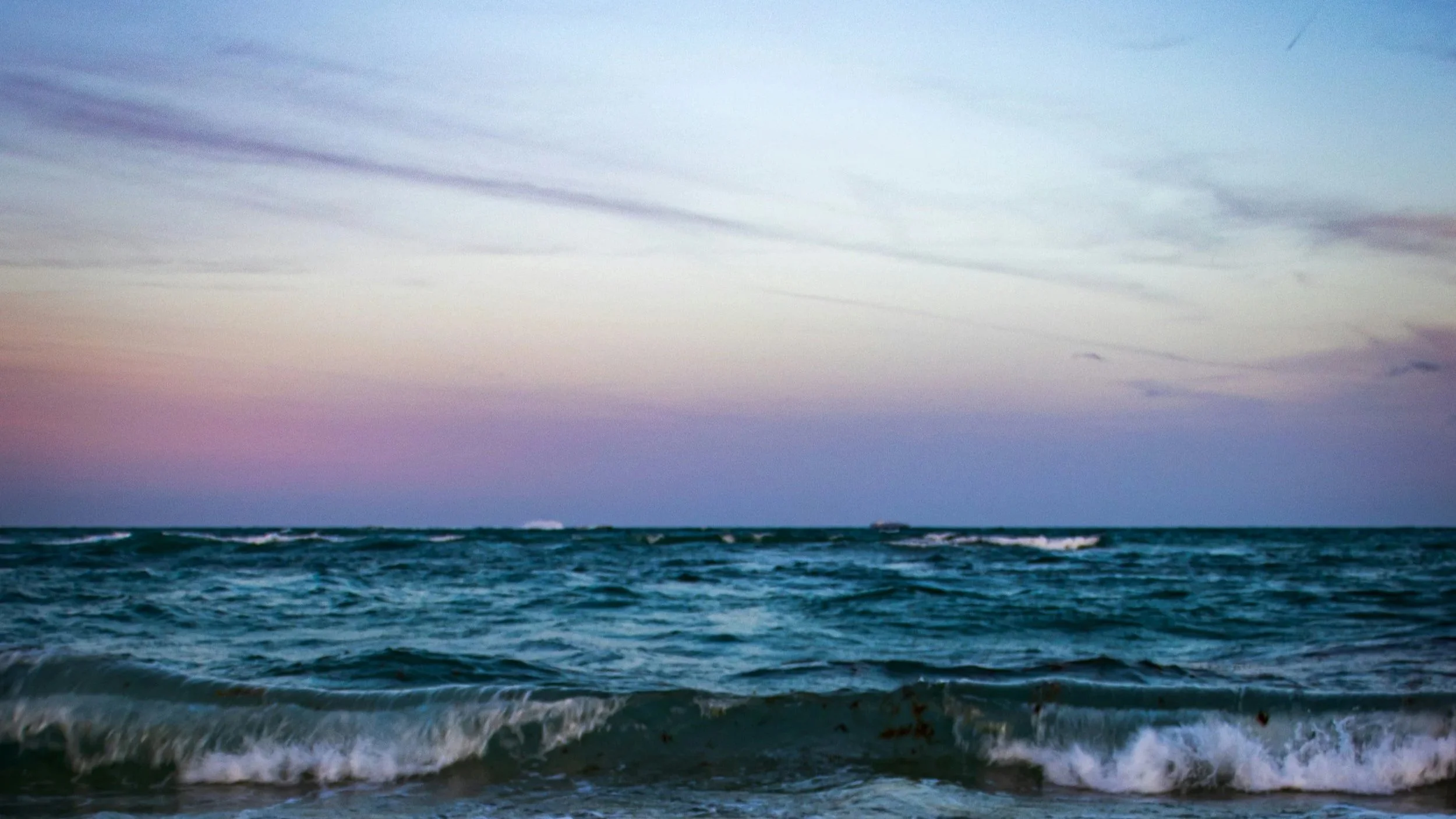 Ocean waves gently crashing on the shore with a sky at sunset showing pink, purple, and blue hues and scattered clouds.