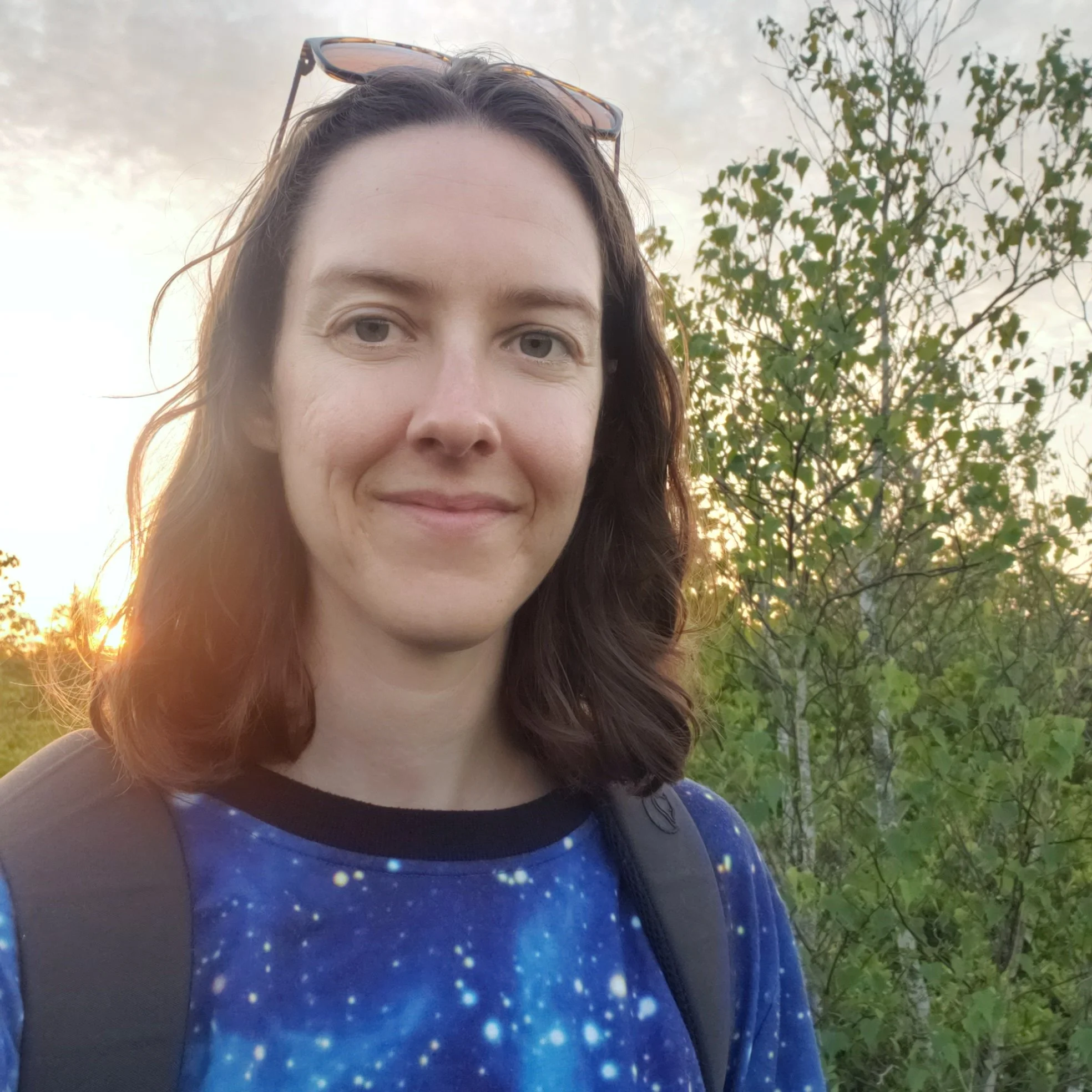Moonlit Tide Massage:Woman with shoulder-length brown hair wearing a galaxy-patterned shirt, sunglasses on her head, and carrying a backpack, outdoors during sunset with trees in the background.