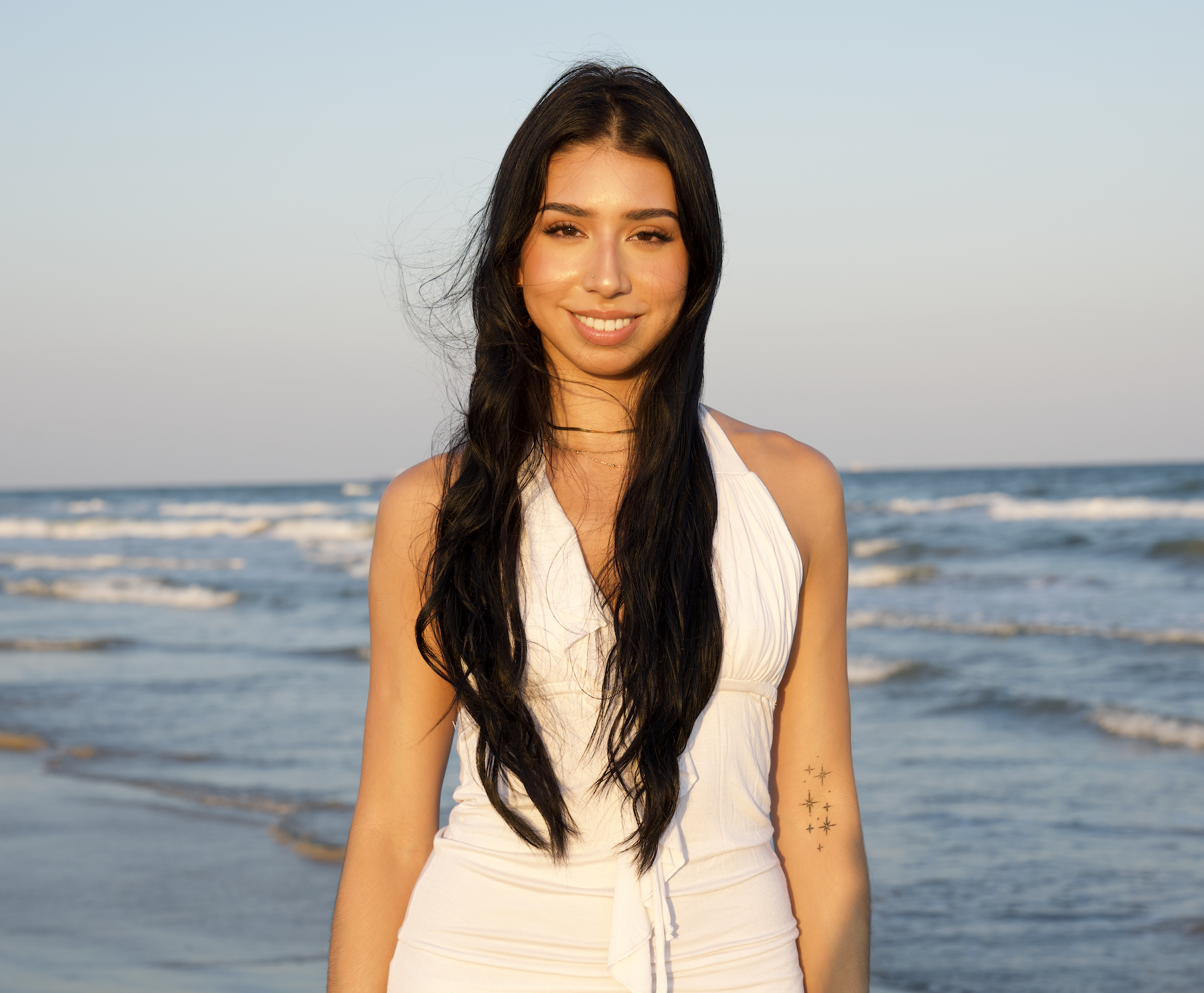 A young woman with long dark hair standing on the beach during sunset, wearing a white sleeveless dress, smiling and looking at the camera, with ocean waves in the background.