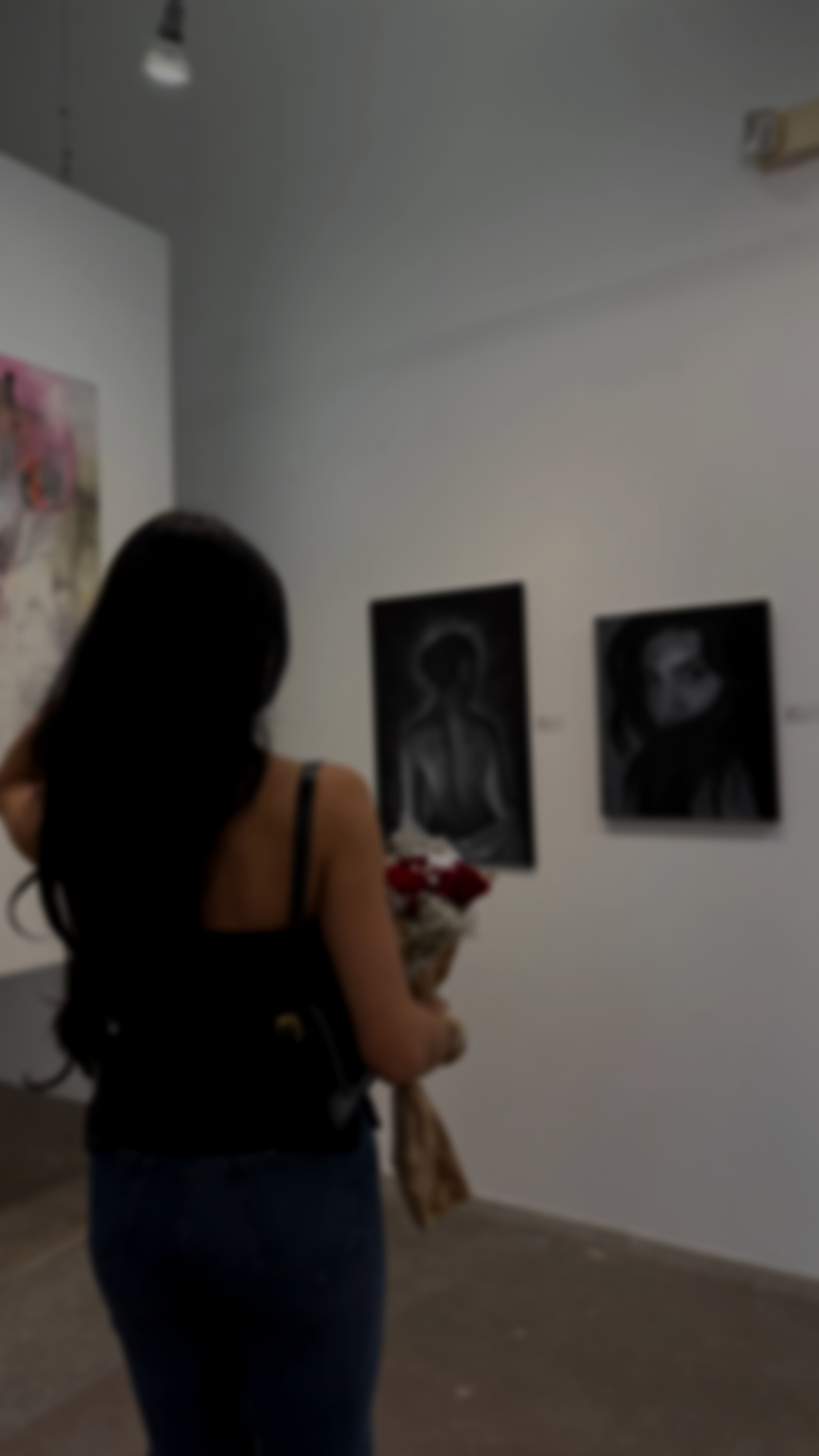 A woman with dark hair wearing a black top and blue jeans standing in an art gallery, viewing black and white portrait paintings on the wall.
