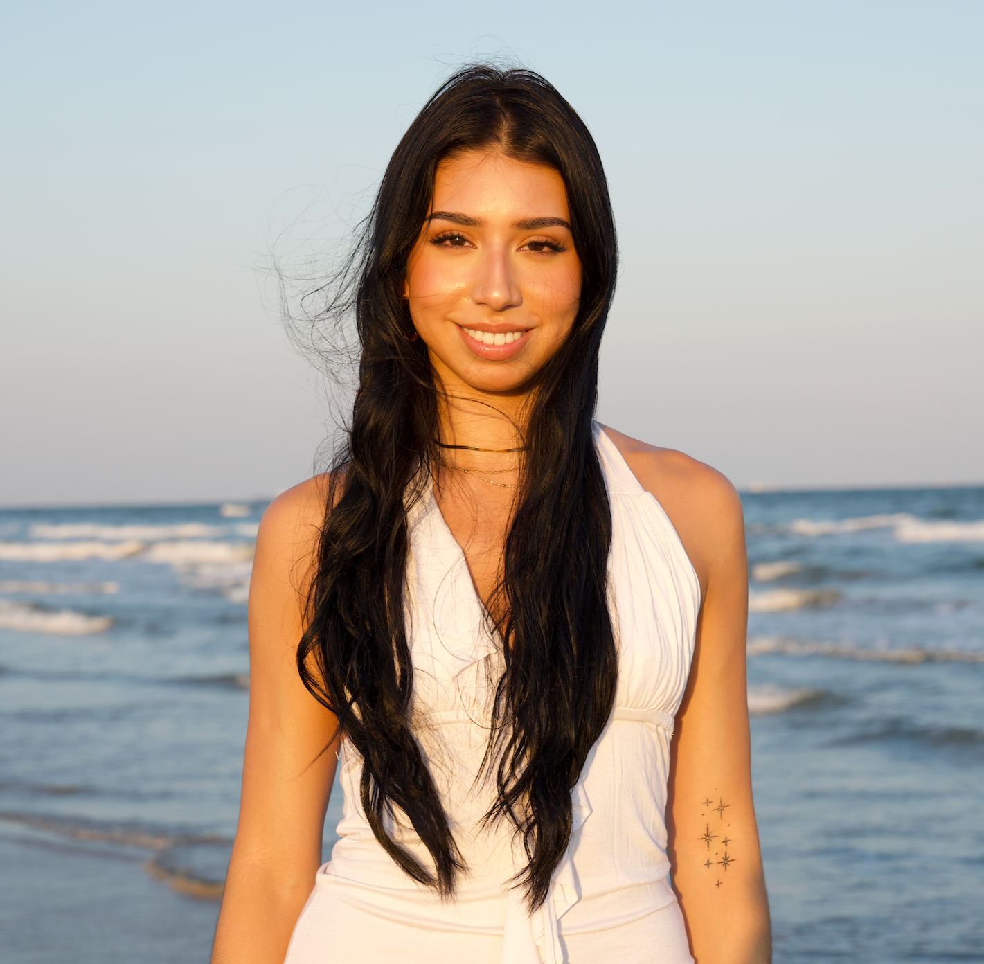 Young woman with long black hair smiling at the beach during sunset, wearing a white sleeveless top, with ocean waves in the background.