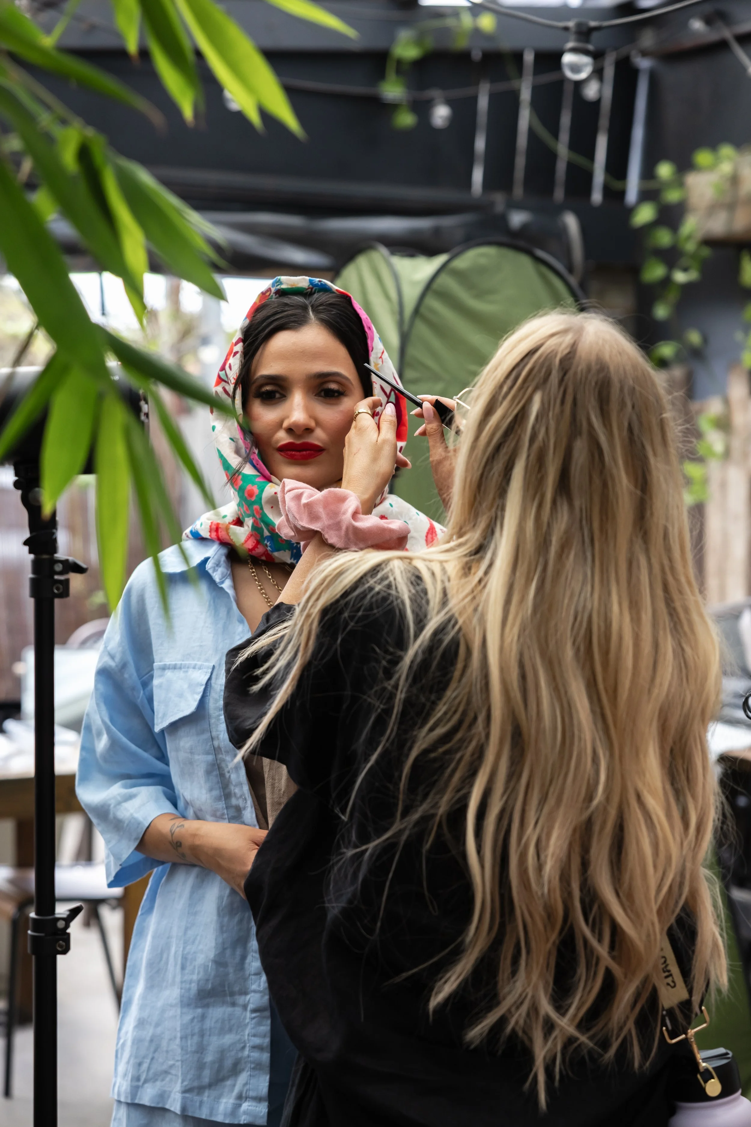 A woman with dark hair and red lipstick is getting her makeup done by a makeup artist with long blonde hair, in an outdoor setting surrounded by greenery, a green tent, and string lights.