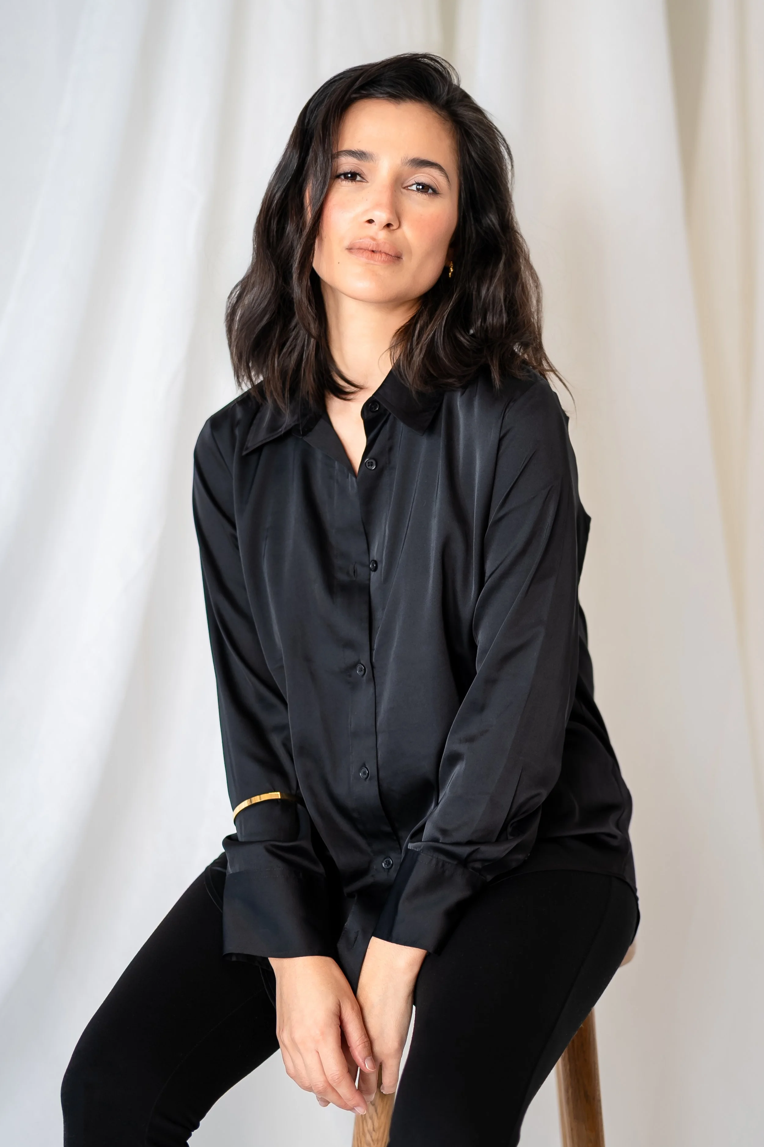 A woman with dark, wavy hair, dressed in a black button-up shirt and black pants, sitting on a wooden stool against a plain white background.
