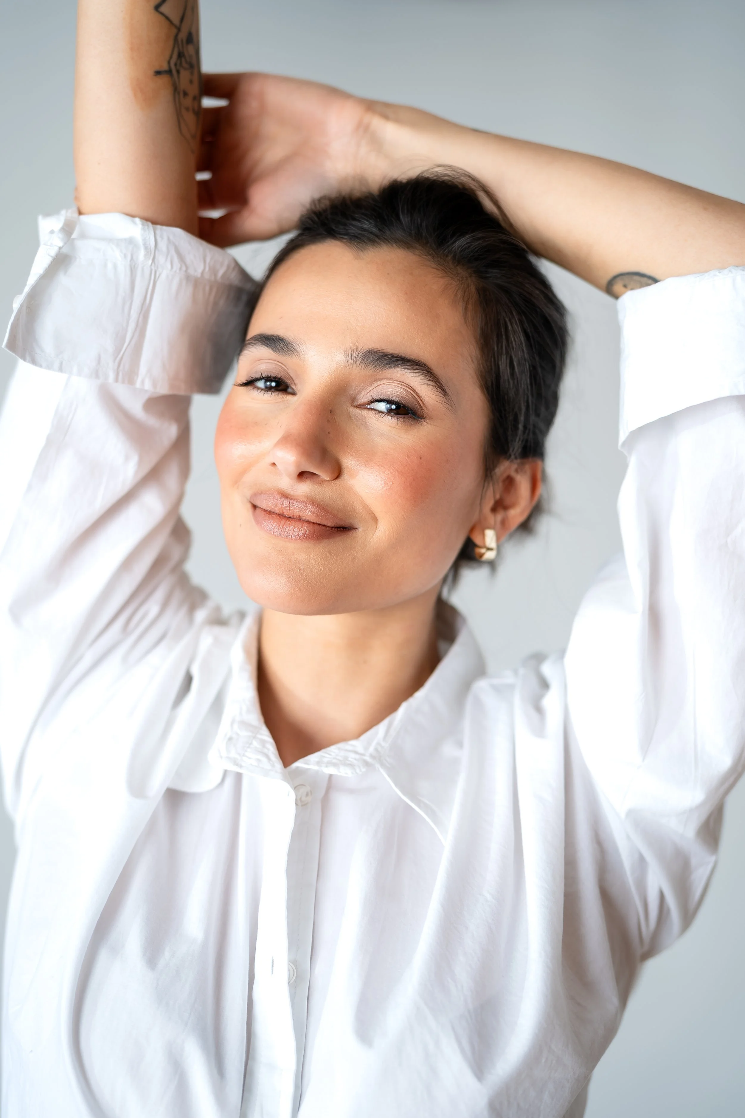 A woman with short dark hair smiling and looking at the camera, wearing a white shirt with rolled-up sleeves and gold earrings, with her arms raised behind her head.