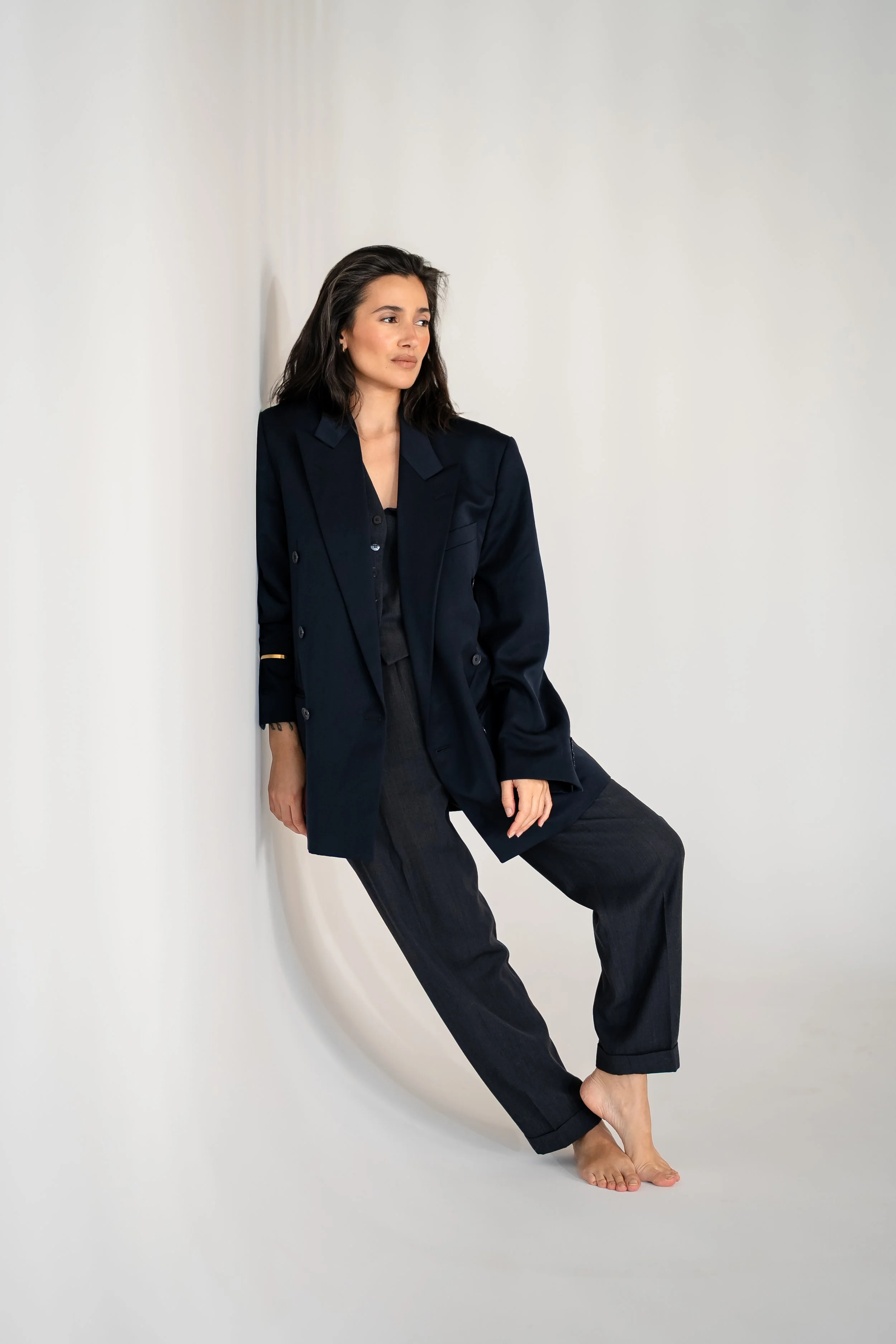 Woman with dark hair in a black blazer and pants, sitting barefoot against a plain white wall.