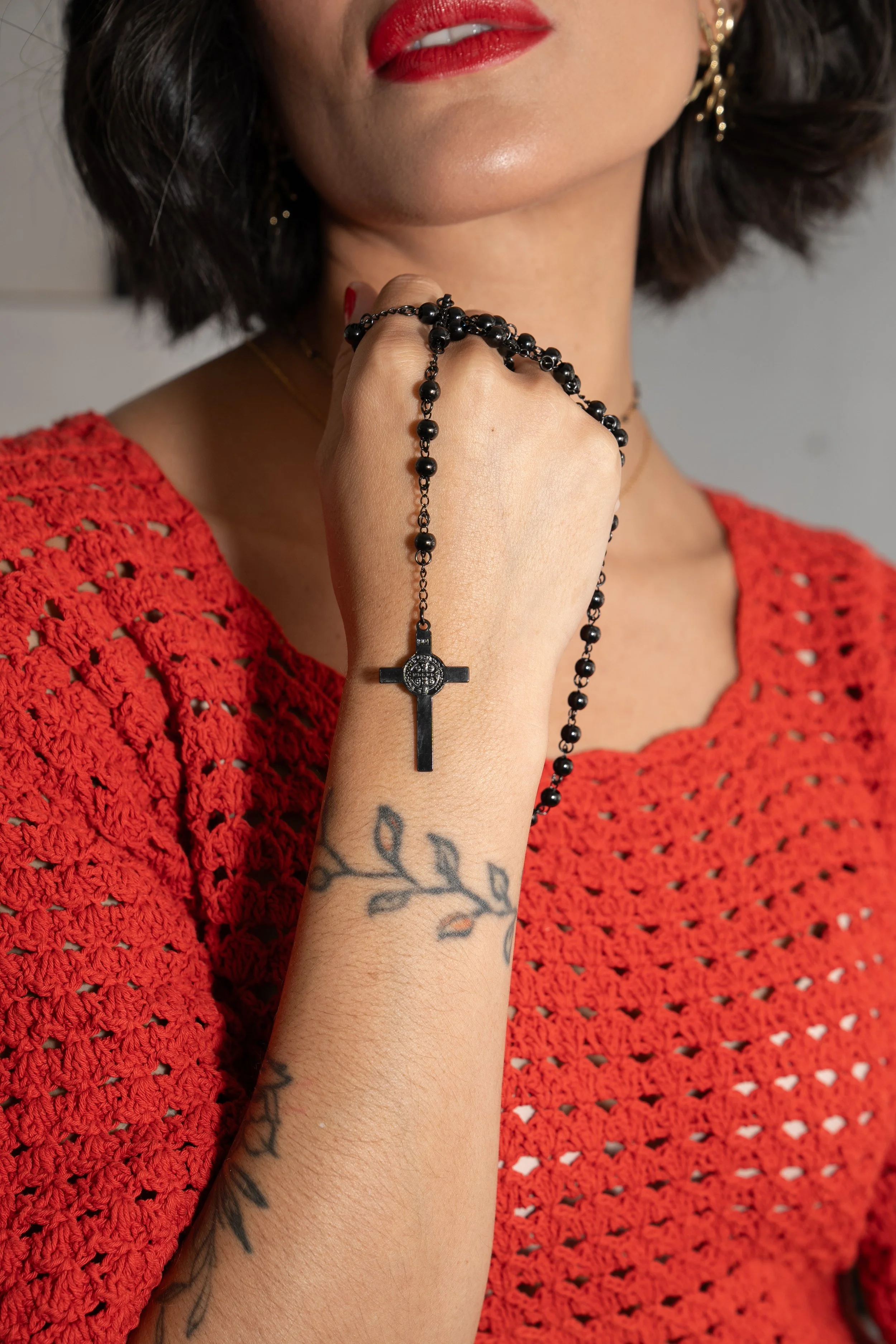 A woman with dark hair, red lipstick, wearing gold earrings, holding a black rosary with a cross pendant near her face, wearing a red crocheted top, and showing a tattoo of leaves on her forearm.