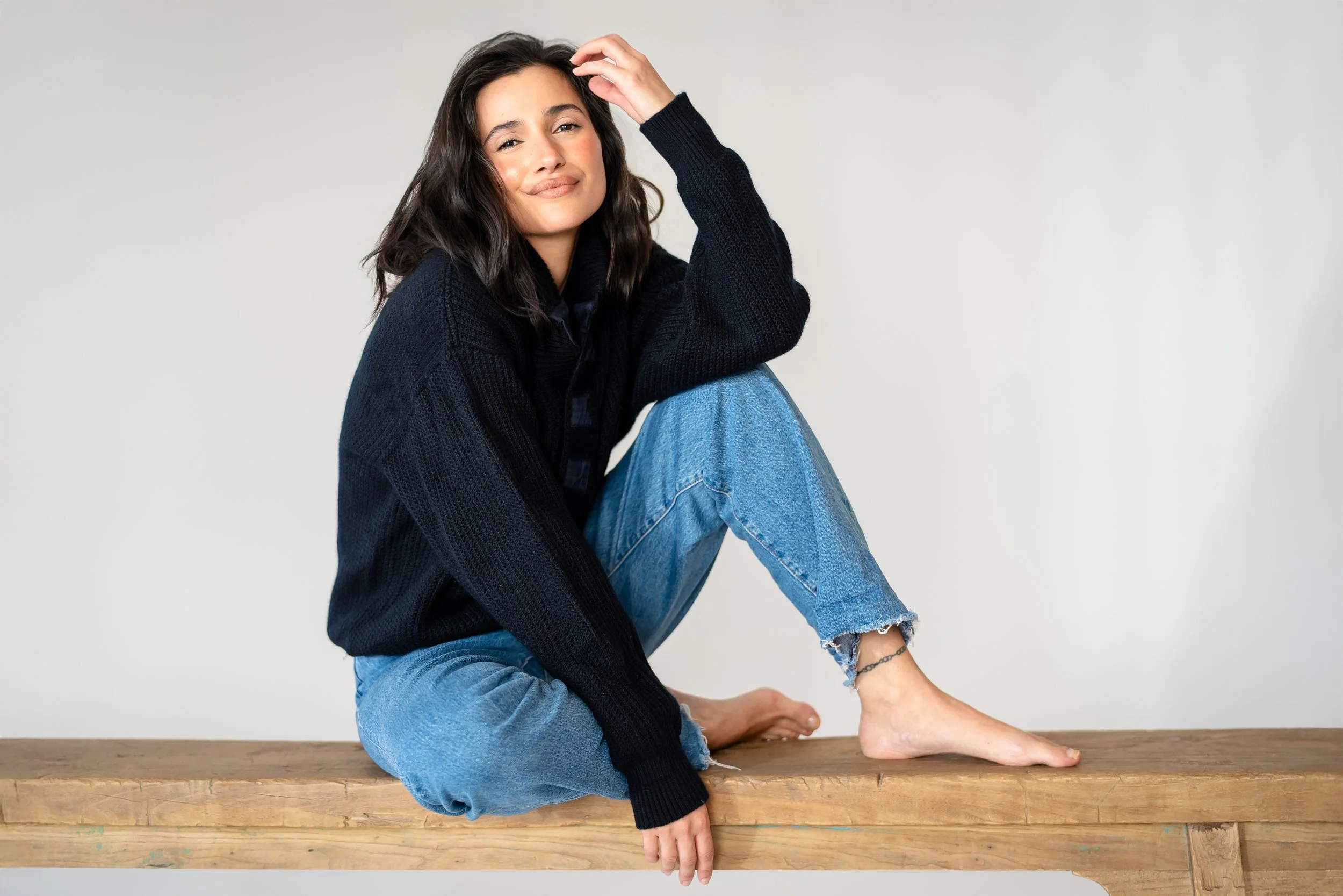 A young woman with dark wavy hair sitting barefoot on a wooden surface, wearing a black sweater and ripped blue jeans, smiling at the camera.